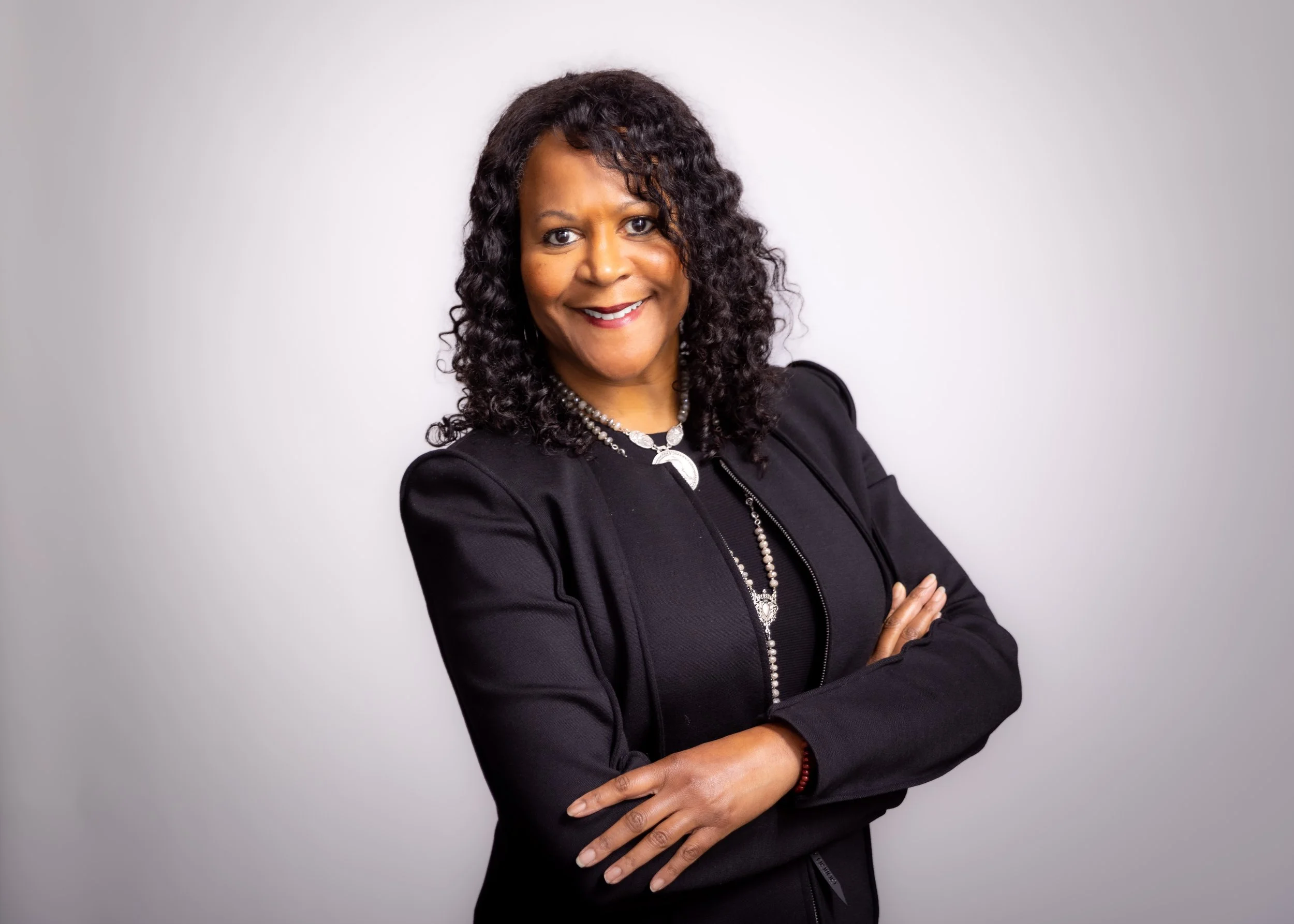A woman with curly black hair, wearing a black blazer and necklace, smiling with arms crossed against a plain white background.