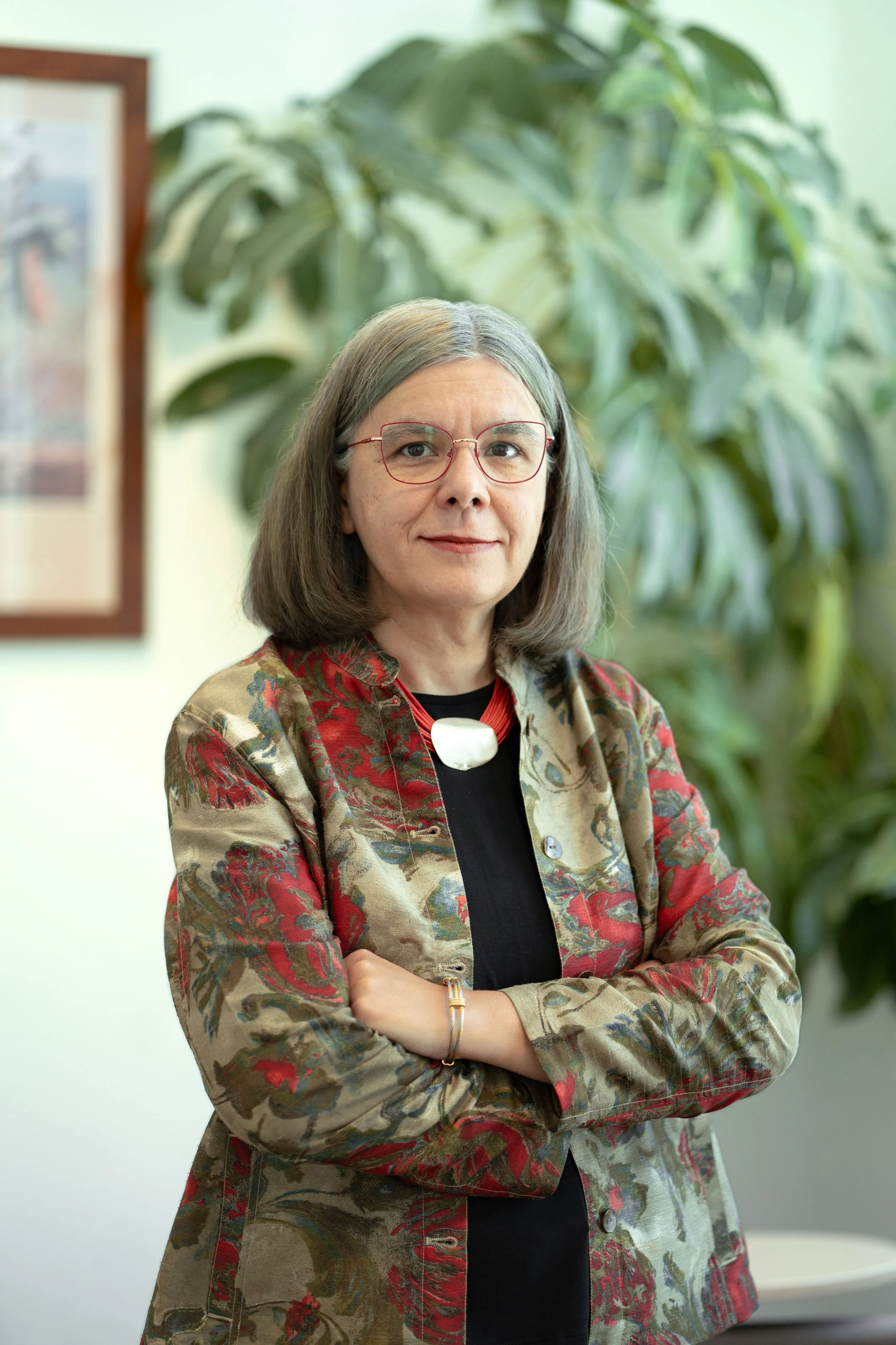 A woman with shoulder-length gray hair and glasses, wearing a patterned blazer and a black top, standing indoors with arms crossed in front of a large green leafy plant and a framed picture on the wall behind her.