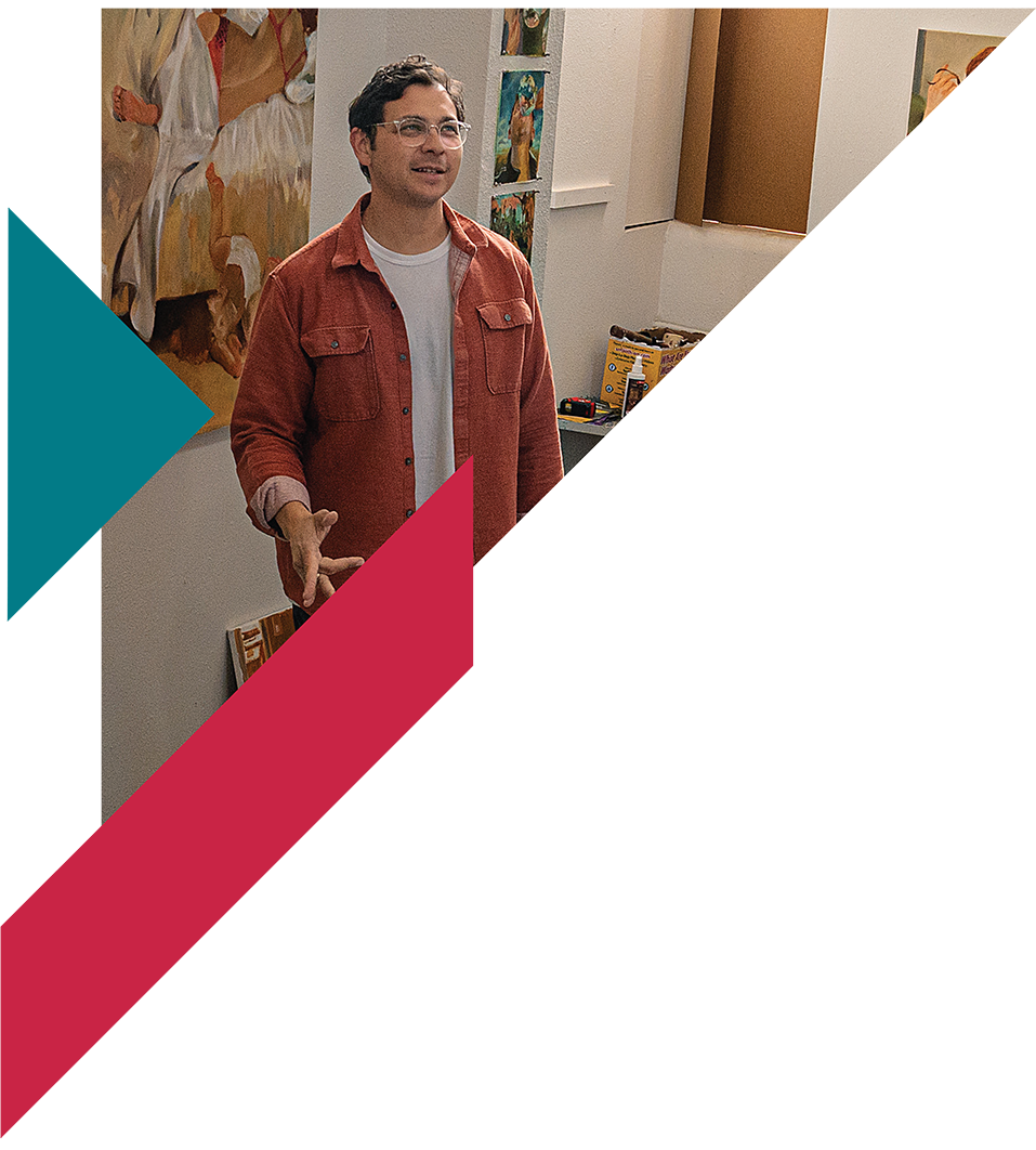 A man with dark hair, glasses, wearing a red shirt over a white t-shirt, standing indoors near artwork and art supplies.