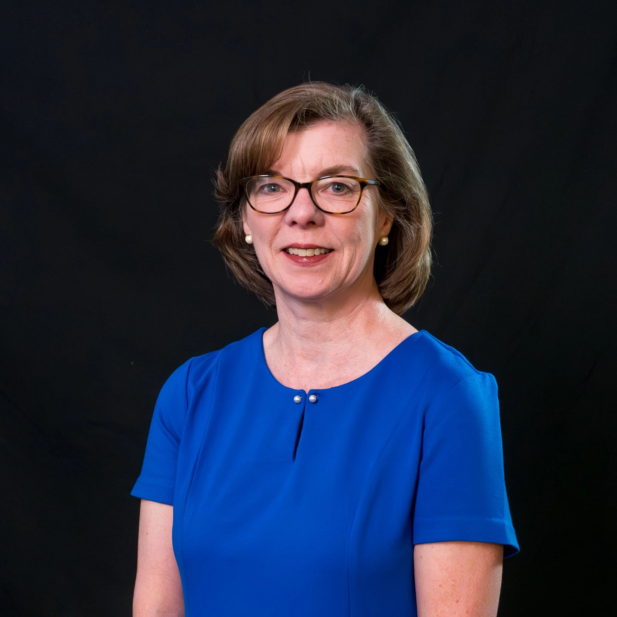A woman with short light brown hair, glasses, and pearl earrings wearing a blue dress with a pearl button. She is smiling against a plain black background.