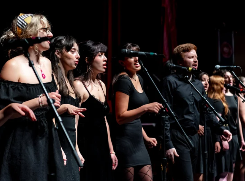 Group of singers performing on stage with microphones, women in black dresses, one woman wearing glasses and a colorful headscarf.