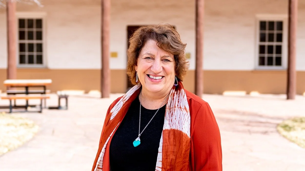 A woman with short curly hair smiling outdoors in front of a building with porches. She is wearing a red jacket, black top, colorful scarf, and turquoise necklace.