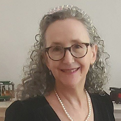 A woman with curly gray hair and glasses smiling, wearing a pearl necklace and black top.