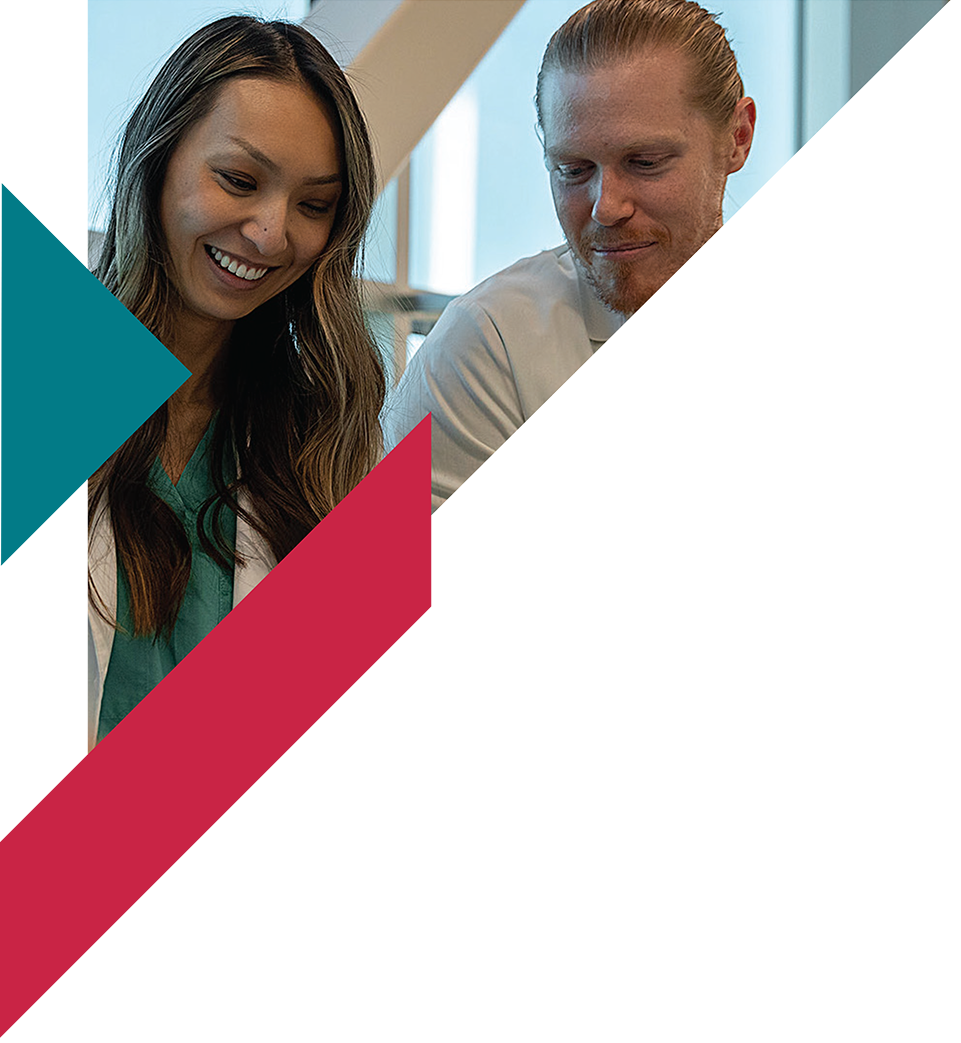 Two smiling healthcare professionals, a woman and a man, looking down at something, in a well-lit medical or office setting.