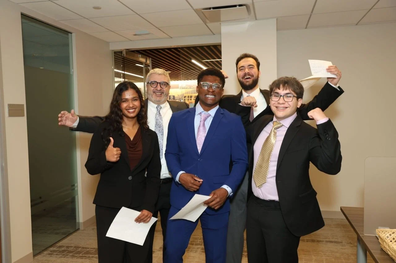 Five diverse professionals in business attire celebrating and smiling in an office setting, with some holding papers.