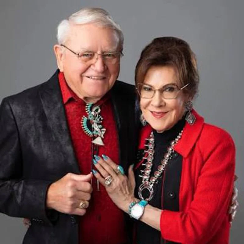 Older man and woman smiling, dressed in red and black with jewelry, standing close together against a gray background.