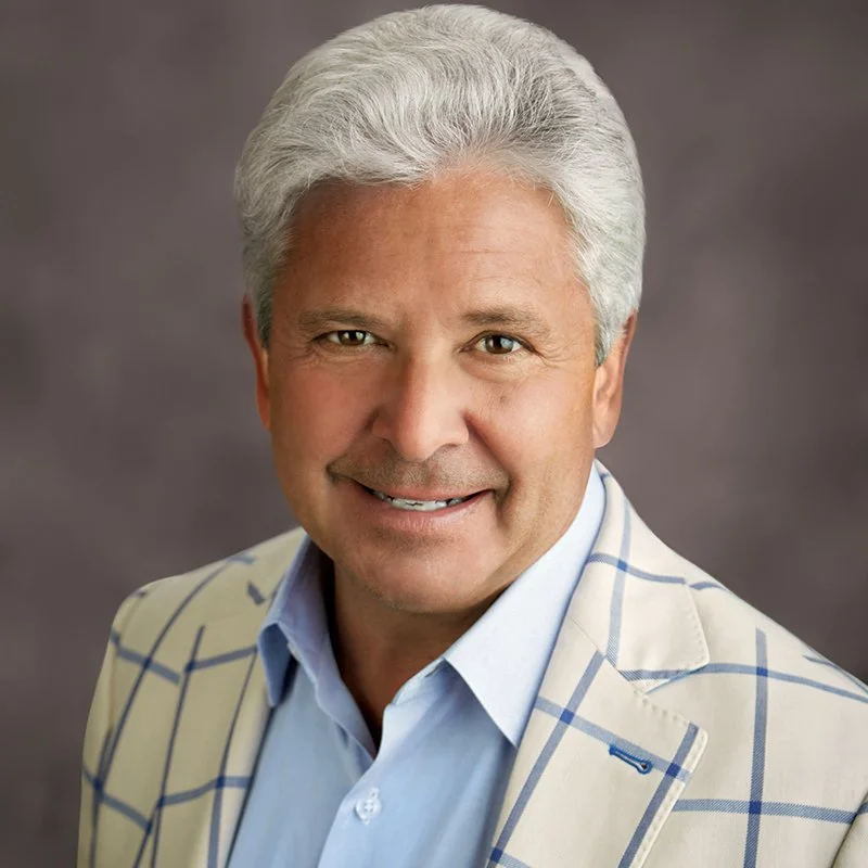 A portrait of a man with gray hair, wearing a light-colored plaid blazer and a light blue shirt, smiling against a gray background.