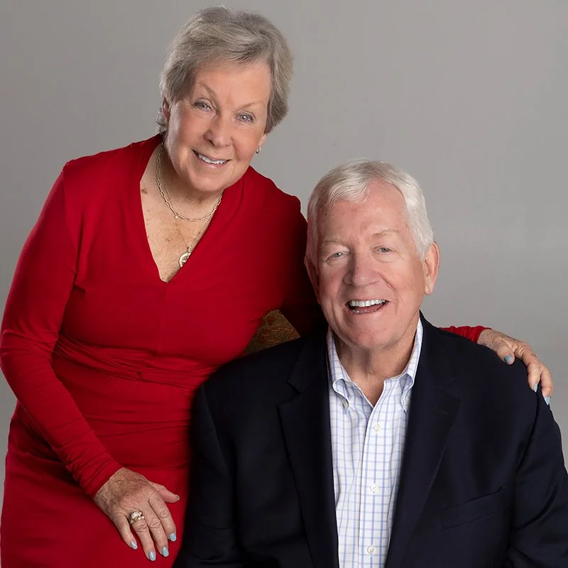 An elderly woman and man smiling for a photo, with the woman standing and resting her hand on the man's shoulder, against a plain gray background.