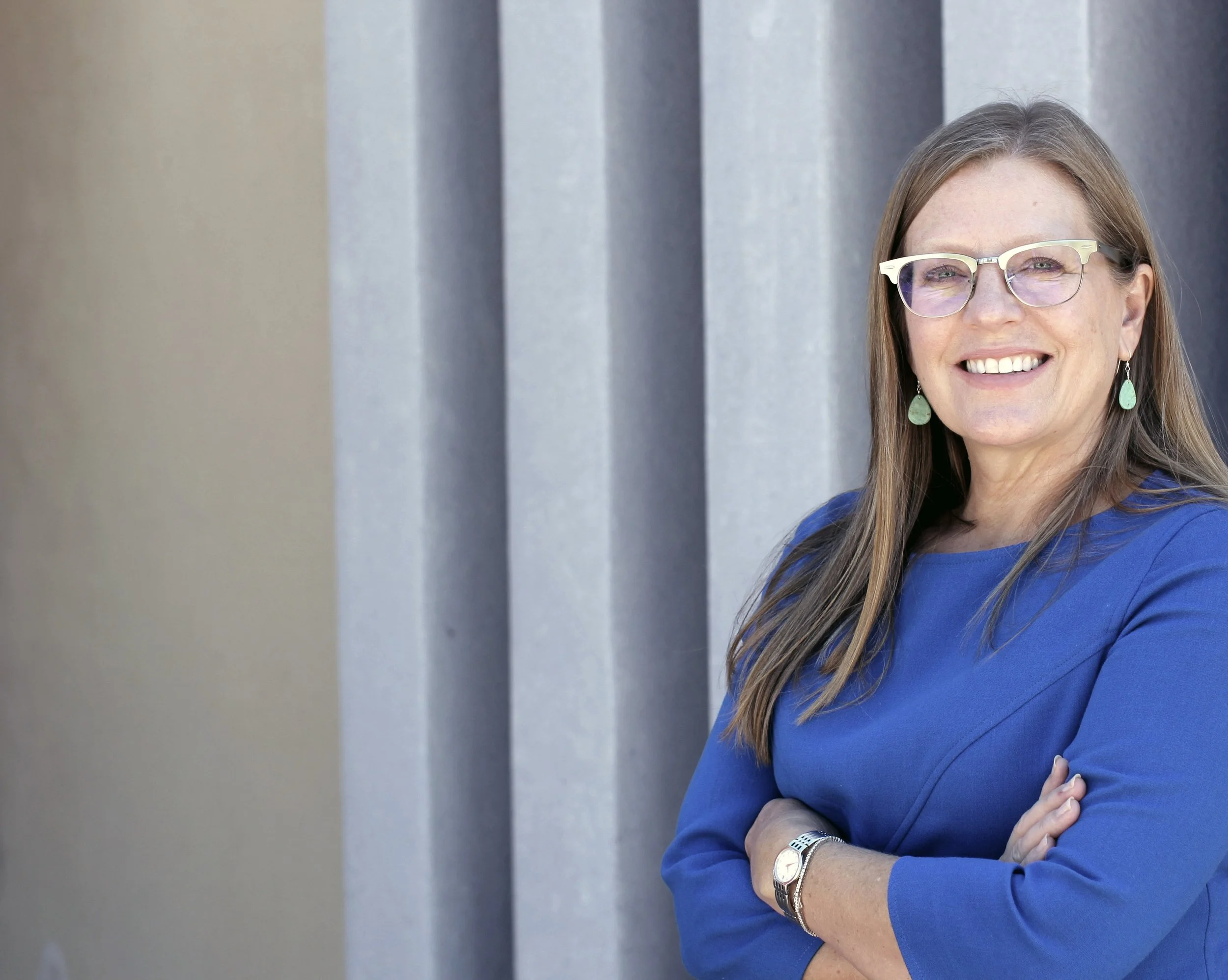 A woman with long brown hair, wearing glasses and turquoise earrings, wearing a blue top, standing outdoors with arms crossed in front of a modern, gray building.
