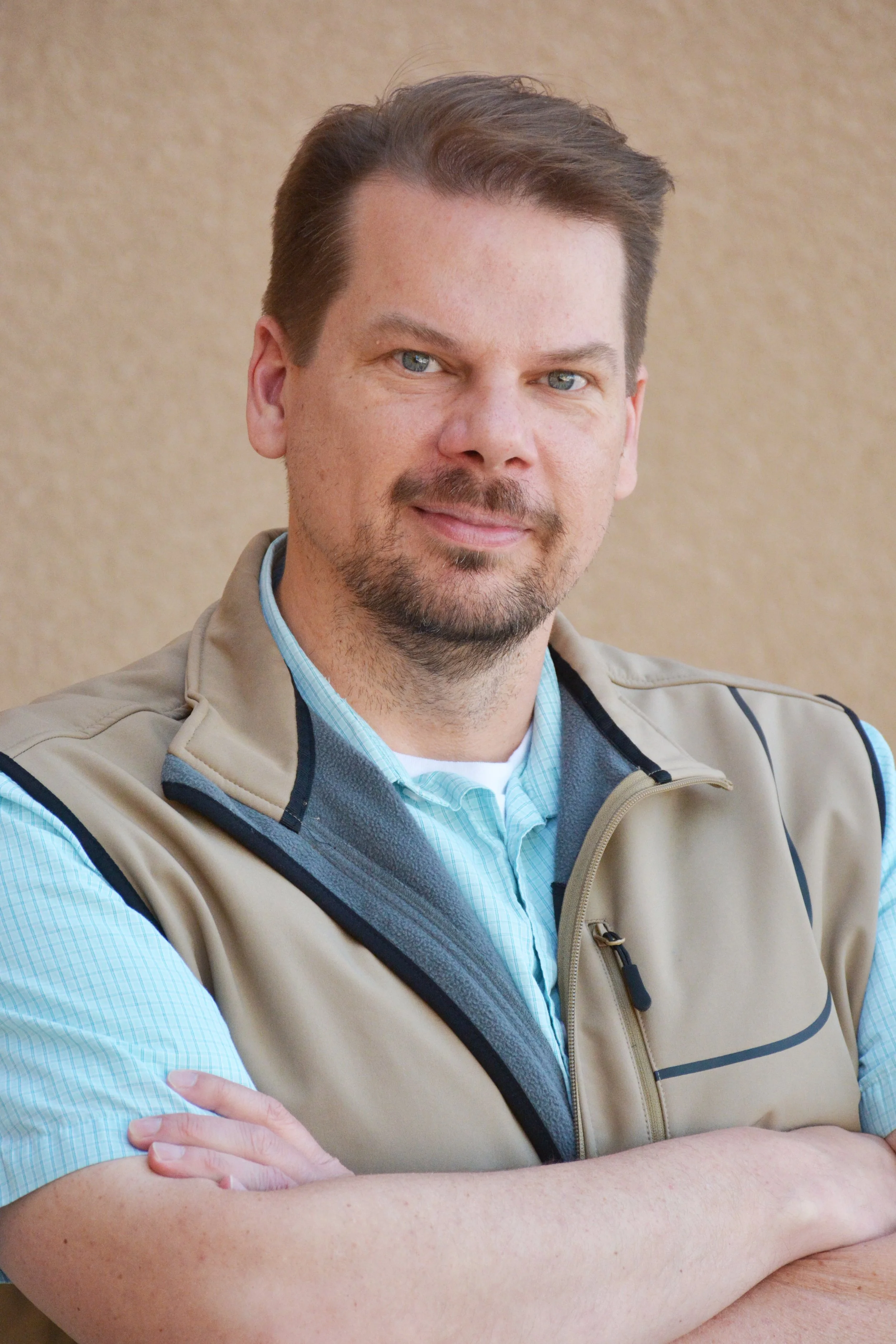 A man with light brown hair and a beard, wearing a tan vest over a blue checkered shirt, sitting with his arms crossed.