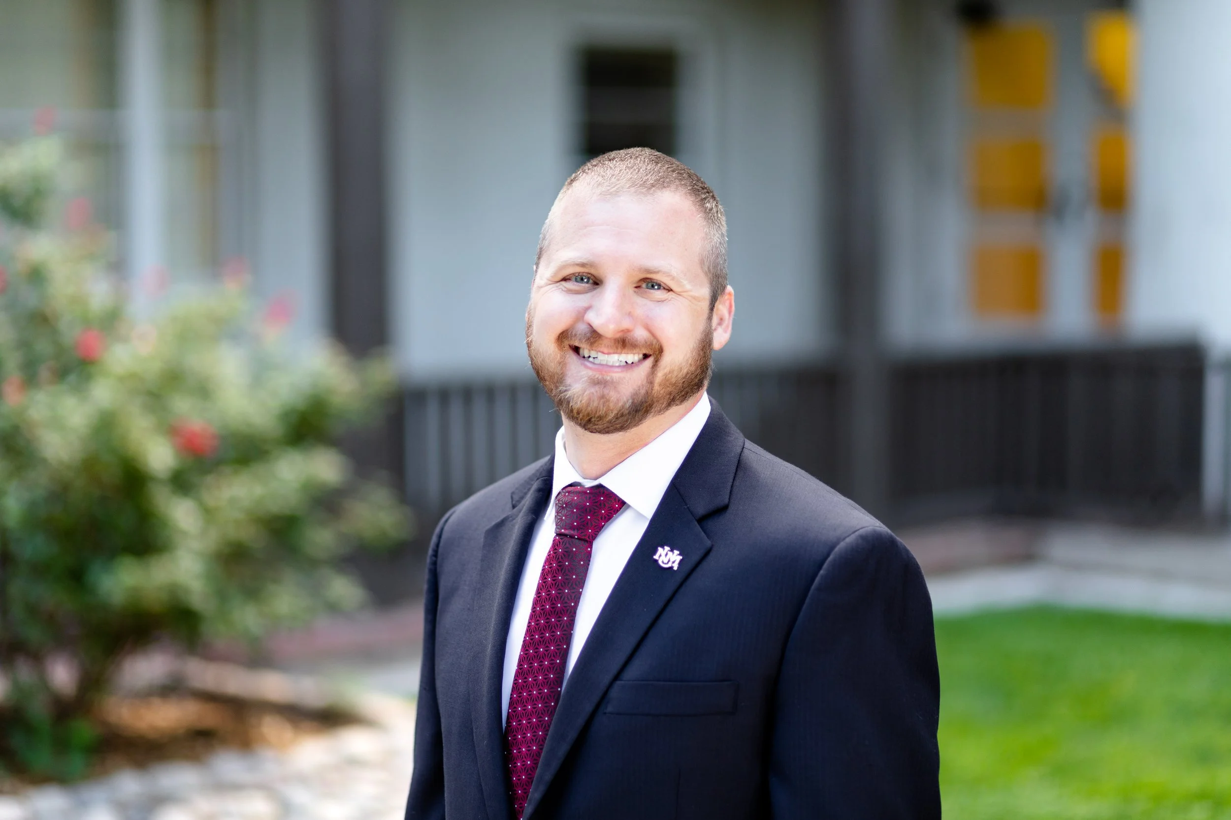 A smiling man in a black suit jacket, white shirt, and maroon tie stands outdoors with a blurry background of a house and greenery.
