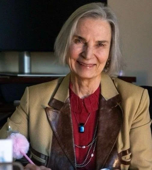 An elderly woman with short gray hair smiling at the camera, wearing a tan blazer over a red top, with a necklace and a black and blue pendant, sitting in a room with a dark background.