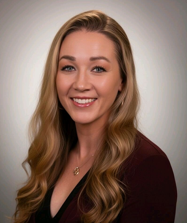 Headshot of a young woman with long, wavy blonde hair, smiling, wearing a dark blazer and a gold necklace, against a neutral background.
