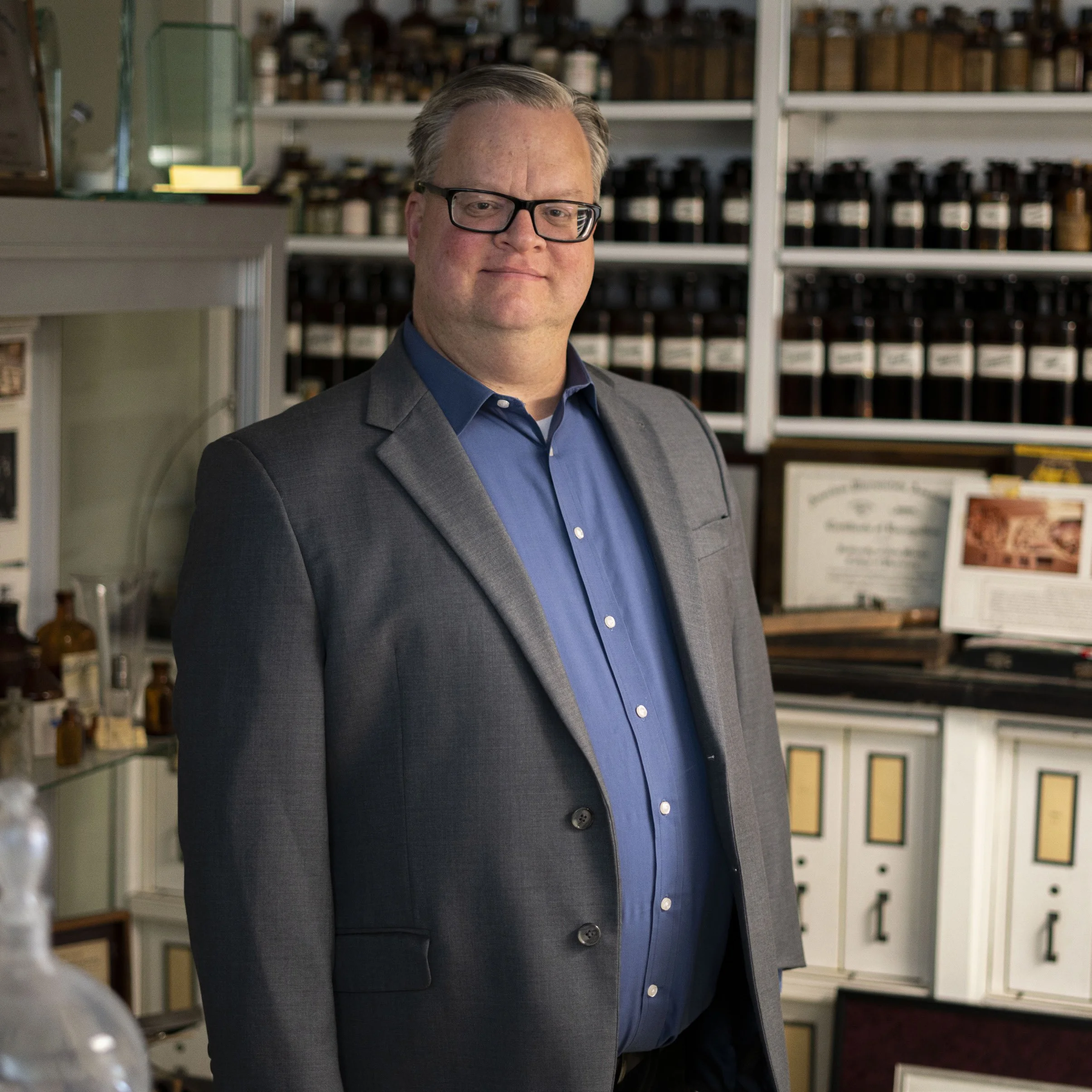 A man with glasses wearing a suit and blue shirt standing in front of shelves filled with bottles and containers in a pharmacy or medical setting.