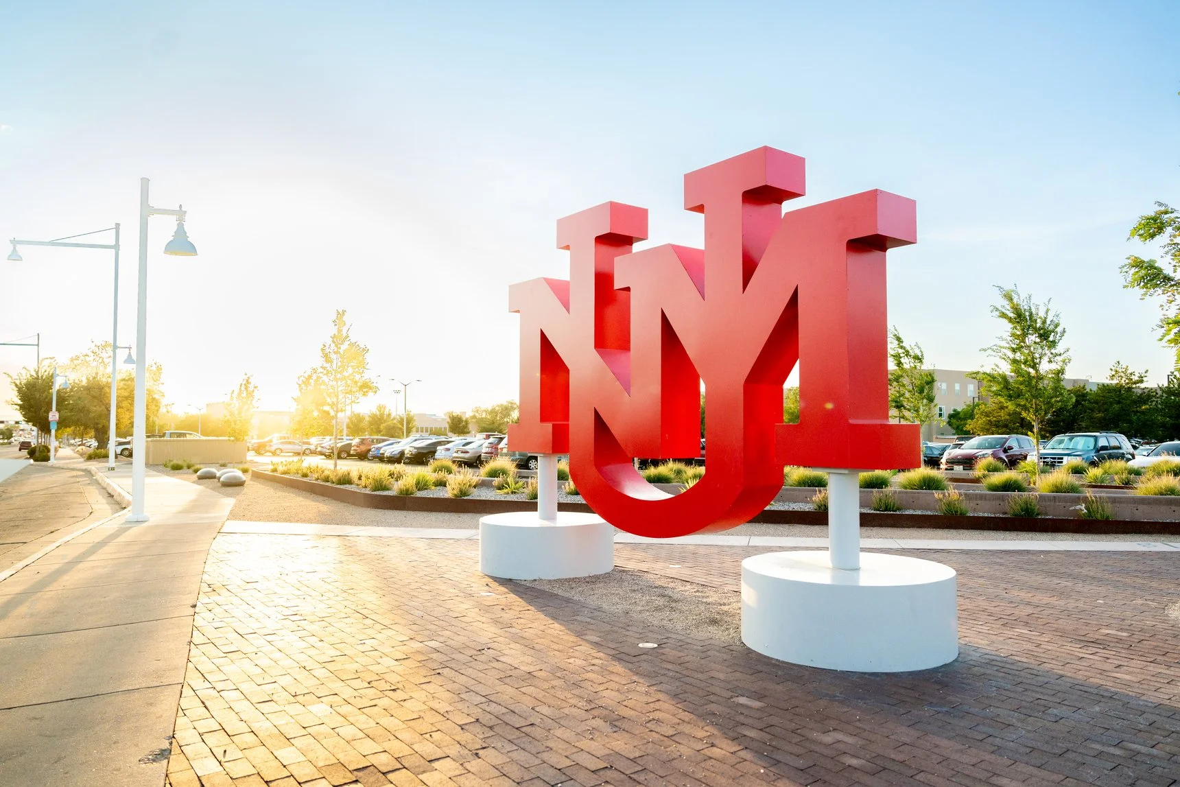Large red NYU sign sculpture outside on a sidewalk with parking lot and trees in the background, sun low in the sky.