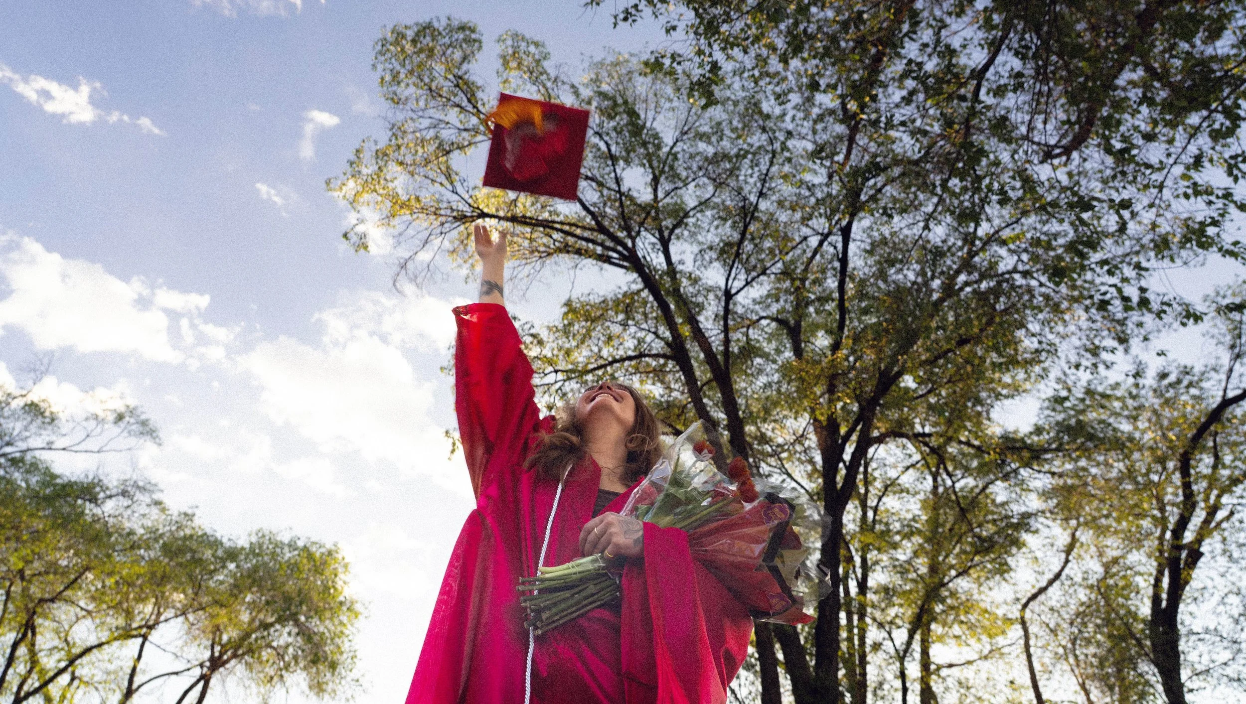 A woman in a pink jacket is smiling and raising a bouquet and a red flag with a yellow star high above her head outdoors, with trees and a partly cloudy sky in the background.