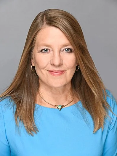 A woman with shoulder-length reddish hair wearing a blue top, smiling at the camera, against a gray background.