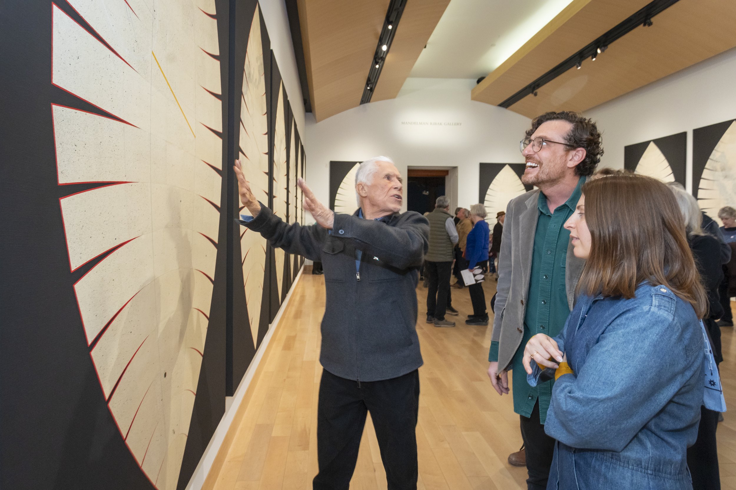An art gallery with several paintings on display and visitors, including a man with glasses and a woman in a denim jacket, observing and discussing the artworks.