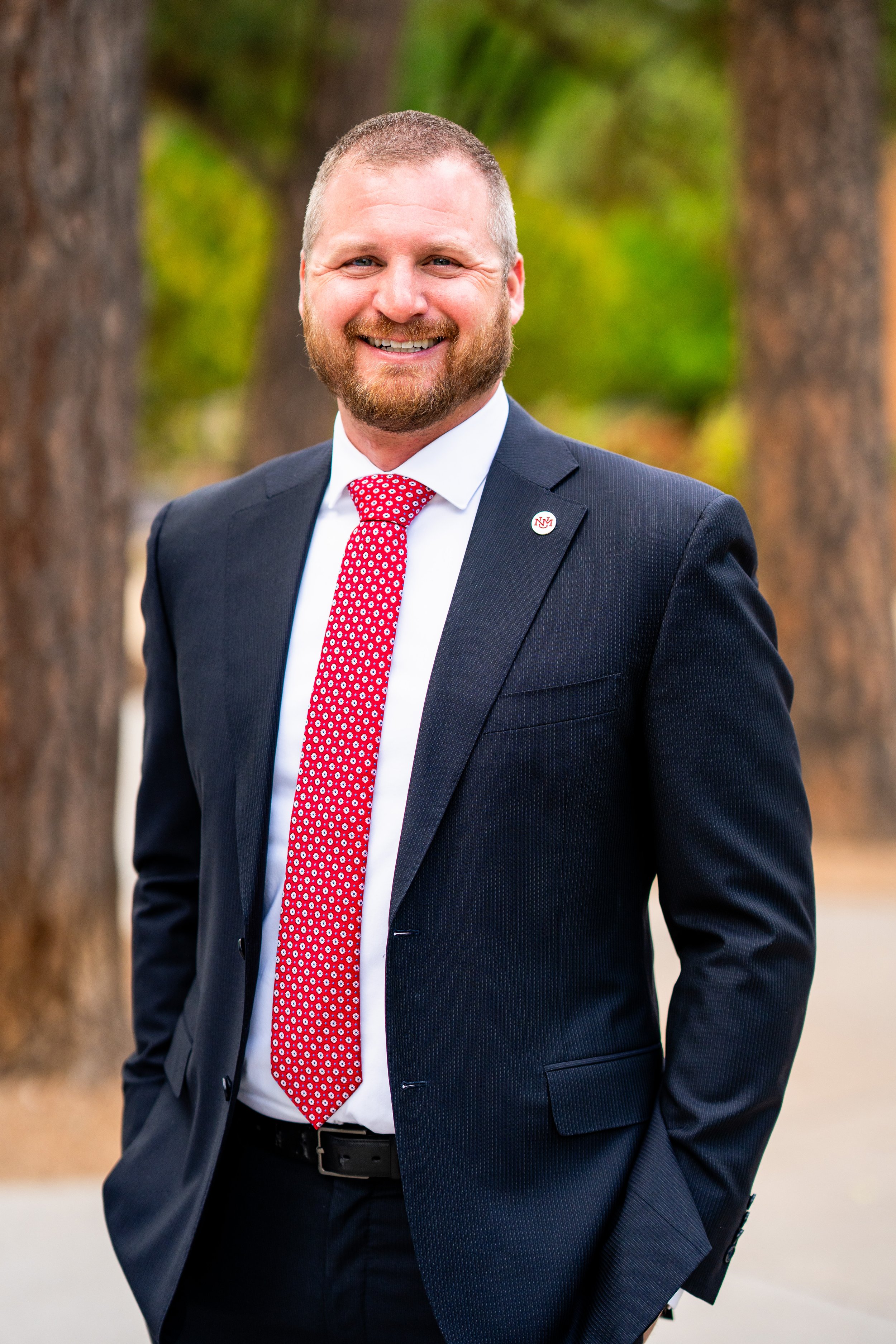 A man in a dark suit and red polka dot tie standing outdoors in front of trees, smiling at the camera.