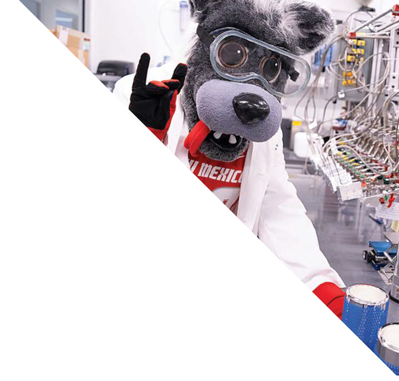 A person dressed in a dog mascot costume with goggles, a hat, and a red tongue is in a laboratory setting with scientific equipment and wiring visible in the background.