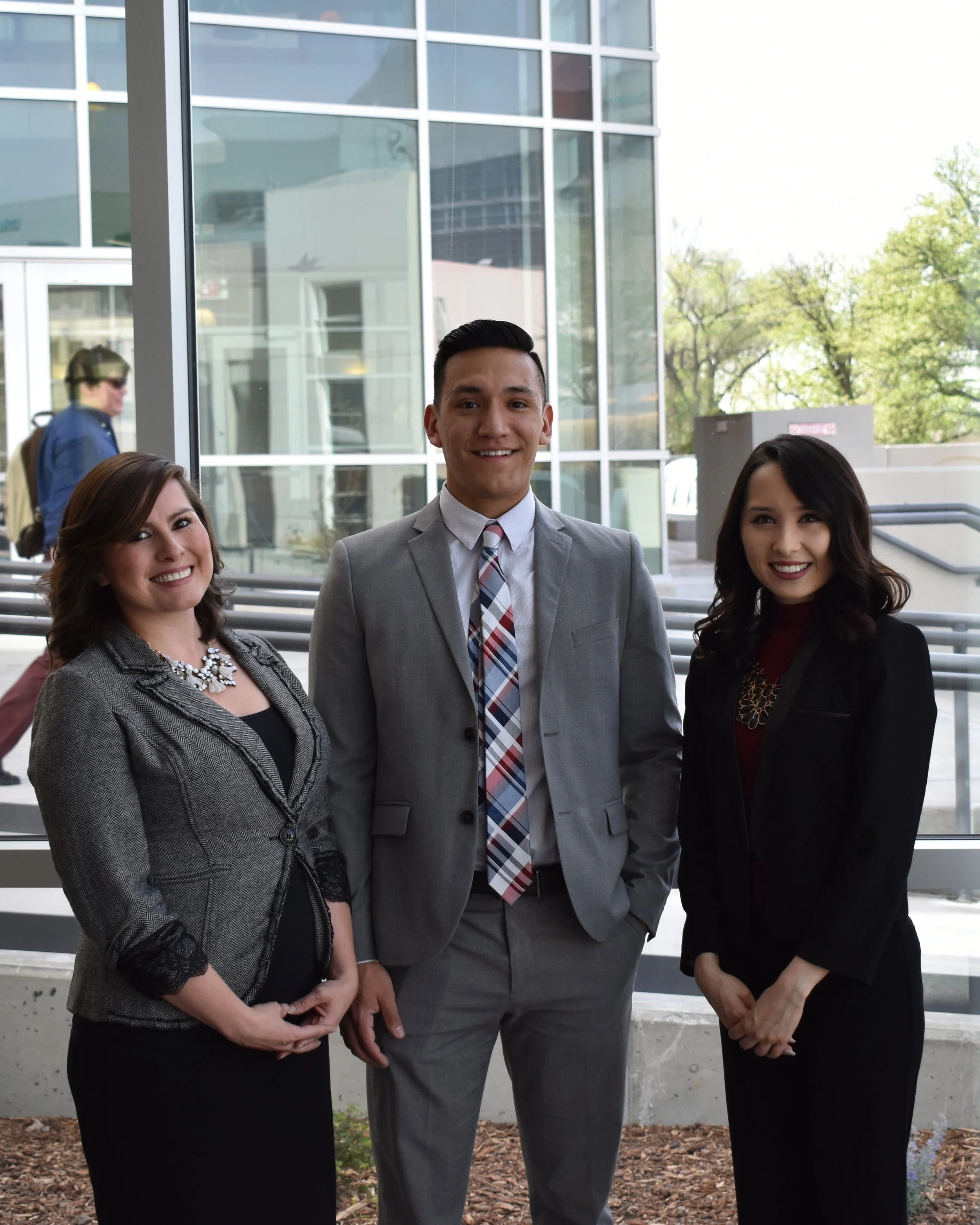 Three professional people, two women and one man, in formal attire standing outdoors in front of a glass building with trees. They are smiling at the camera.