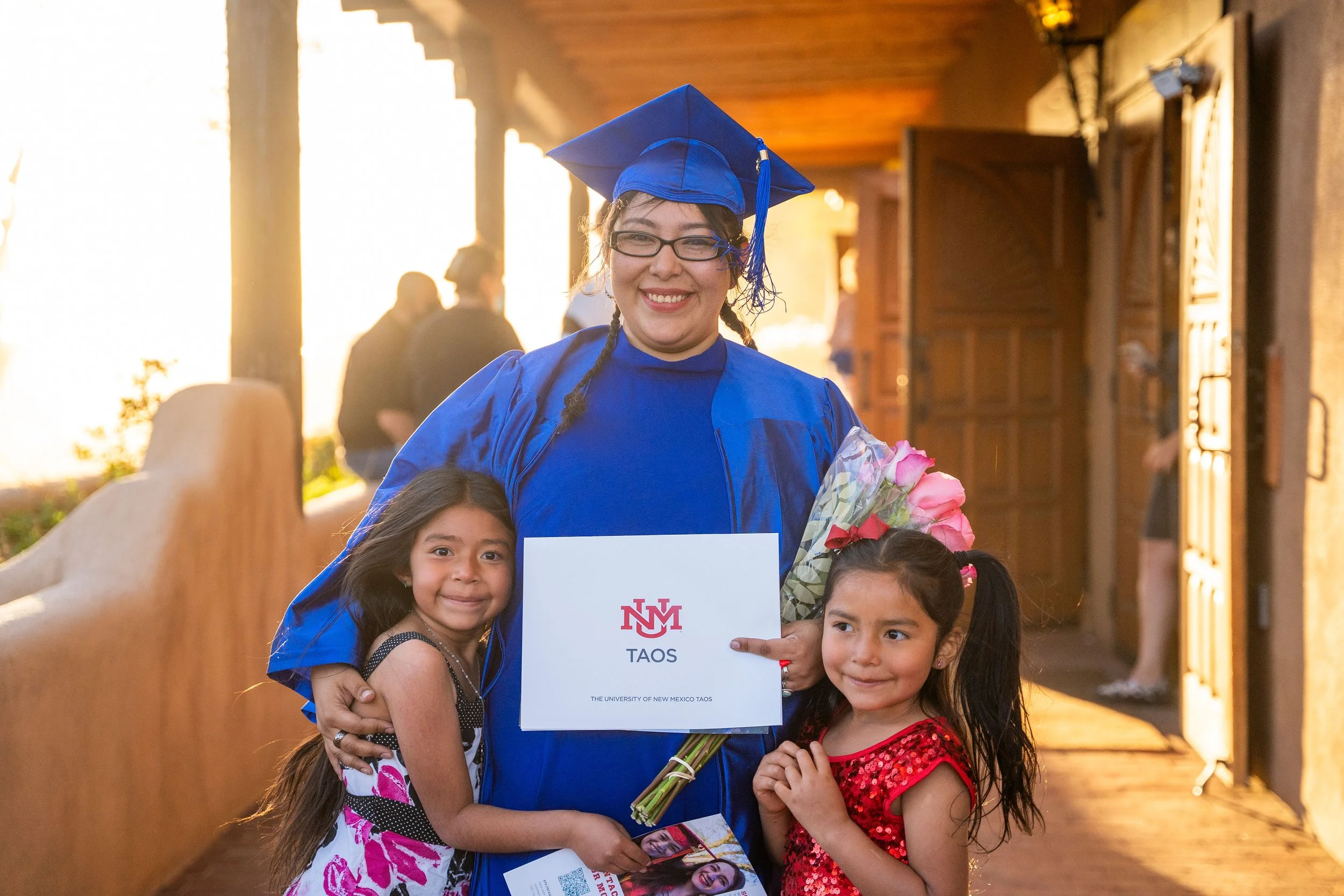 A woman in a blue graduation cap and gown smiling, holding a bouquet of pink roses and a graduation certificate with the logo of the University of New Mexico Taos, surrounded by four young girls. The background shows warm sunlight streaming in through large windows inside a building.