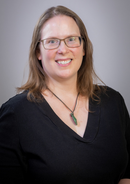 A woman with light skin, shoulder-length light brown hair, glasses, and a black top, smiling at the camera with a gray background.