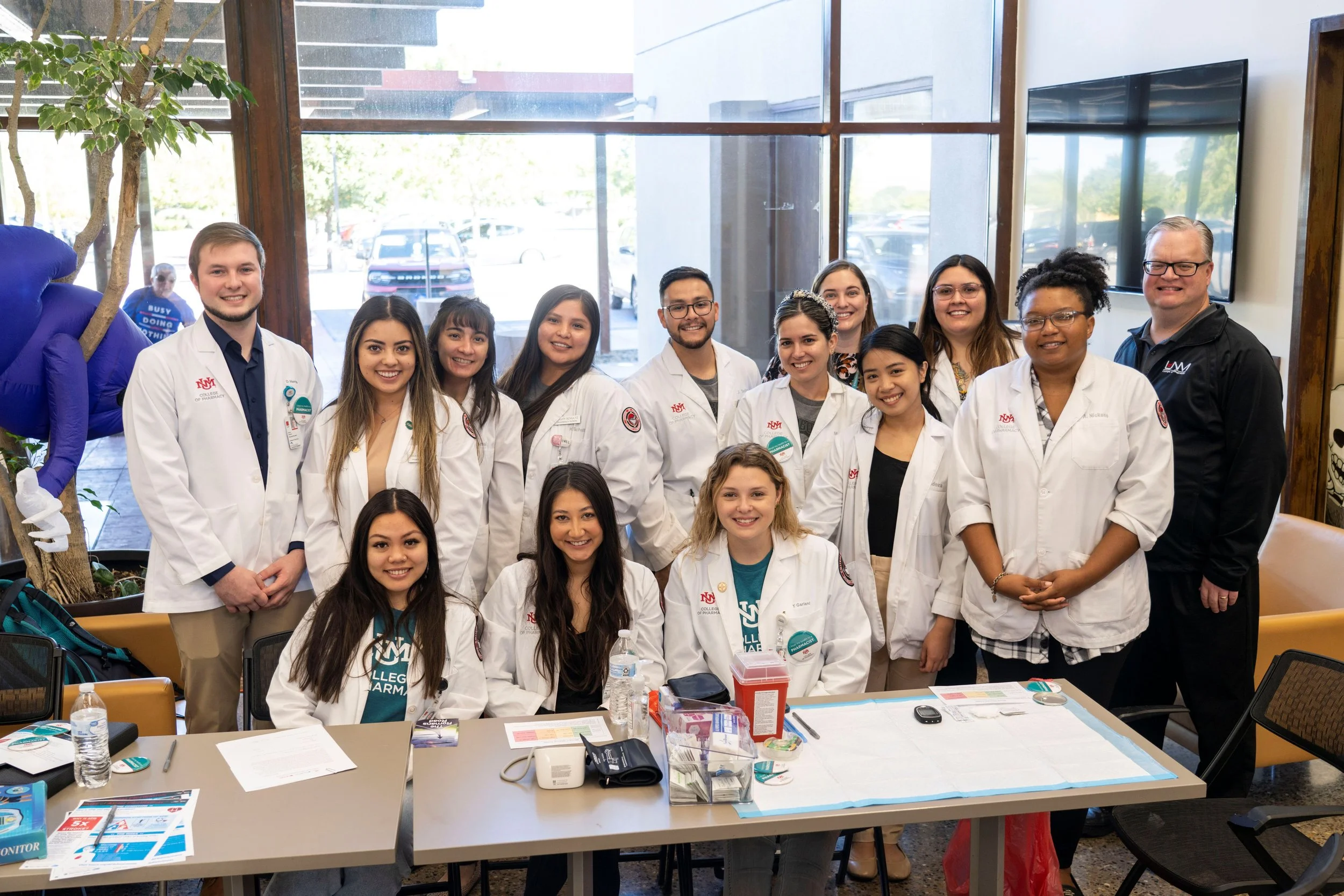Group of diverse students and two adults in medical coats posing for a photo inside a building with large windows. The table in front holds papers, bottled water, and medical supplies, indicating a medical or academic event.