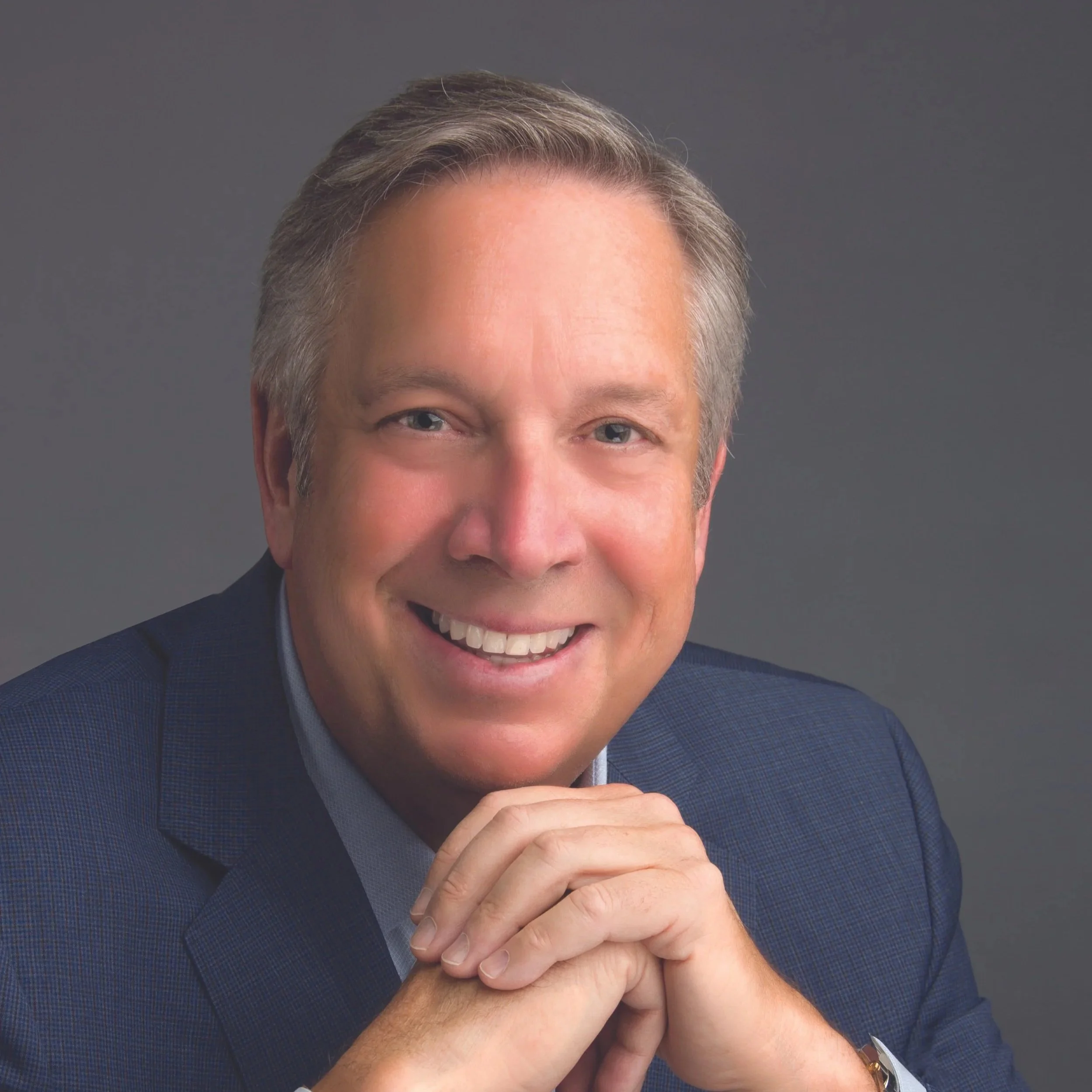 Close-up of a smiling man with gray hair, wearing a dark blue suit, with hands clasped together in front of him, against a gray background.