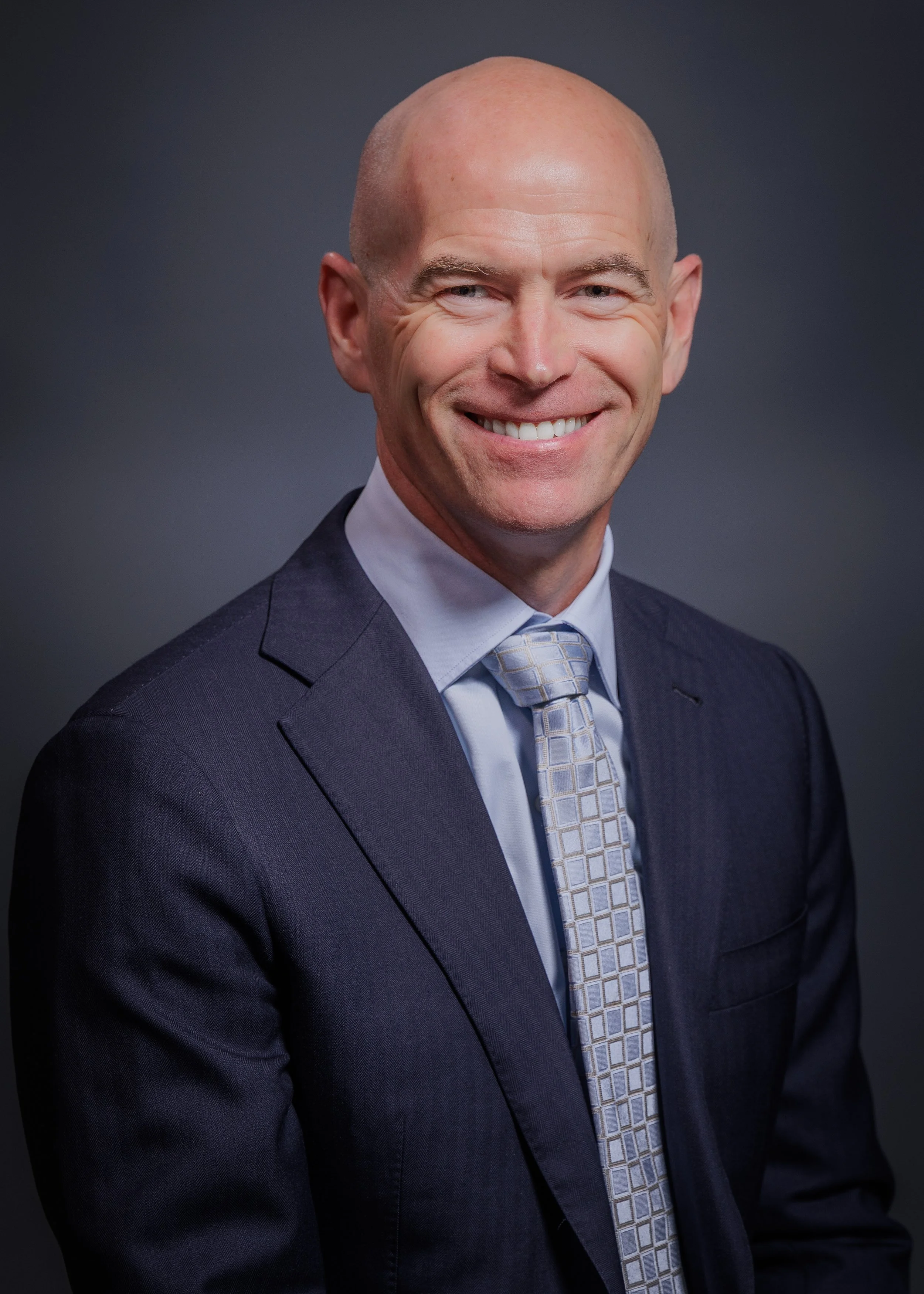 Headshot of a smiling middle-aged man in a suit and tie against a dark background.