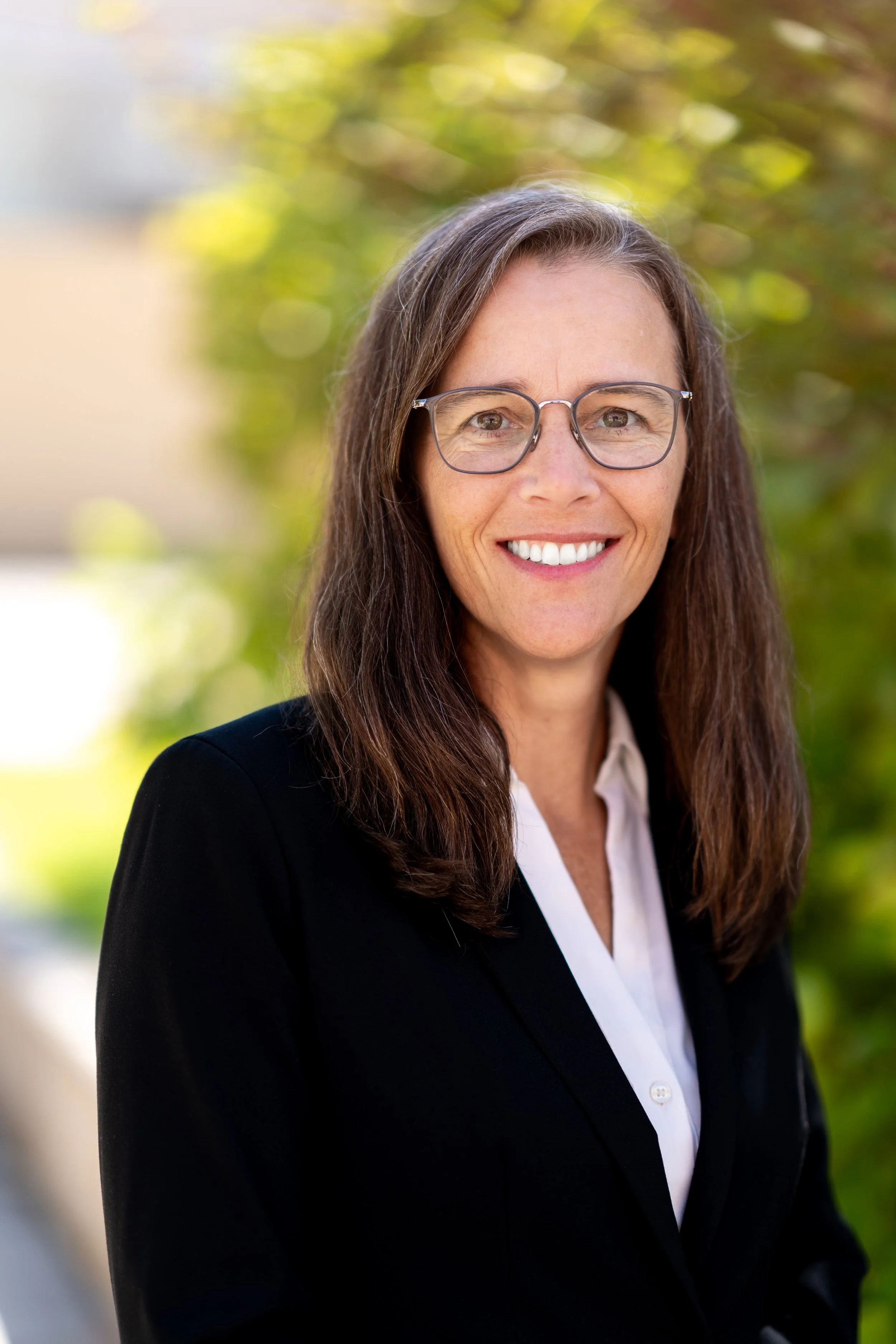 A smiling woman with brown hair and glasses wearing a black blazer and white shirt standing outdoors with green trees in the background.