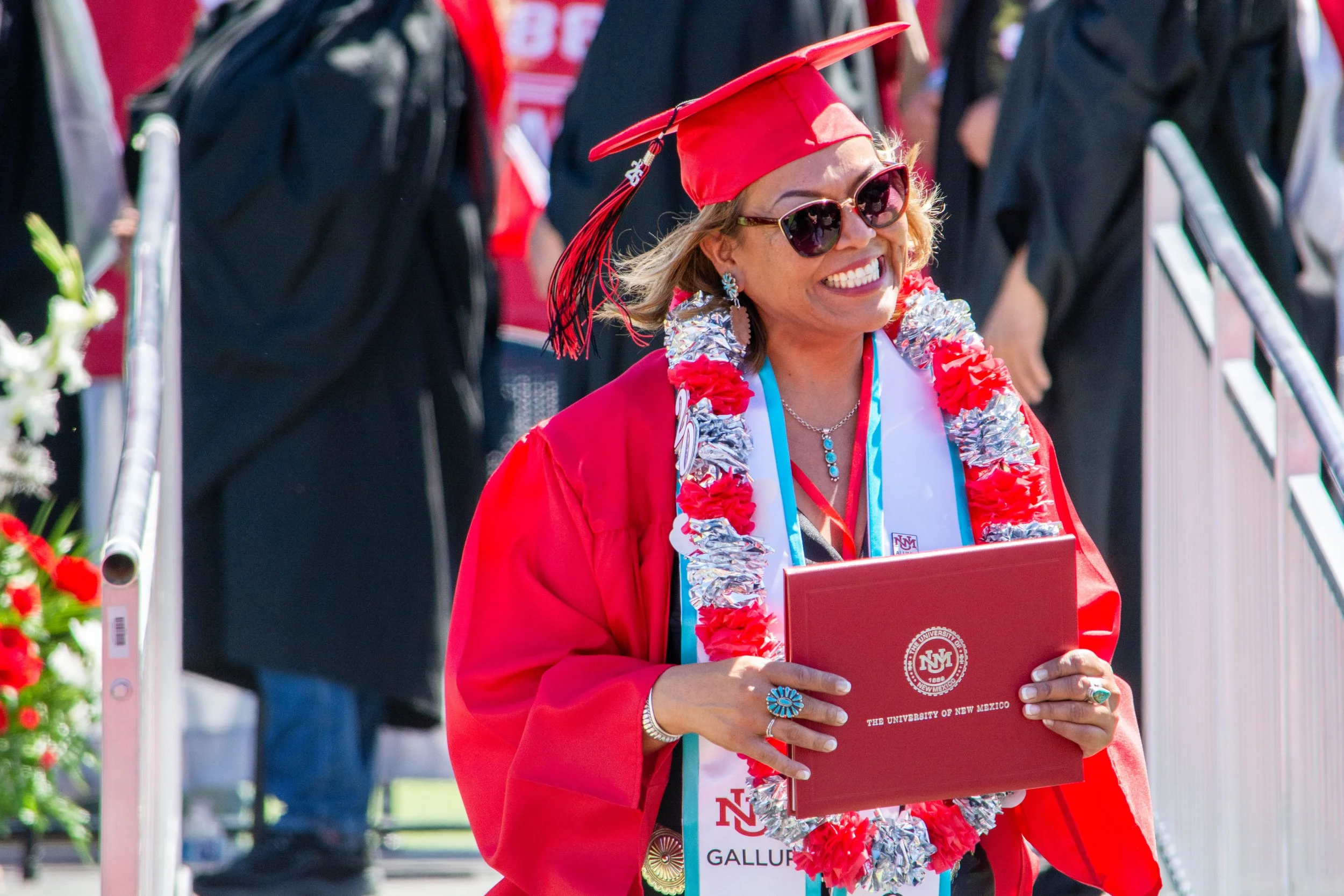 Woman in red graduation gown and cap smiling and holding a diploma at a graduation ceremony.