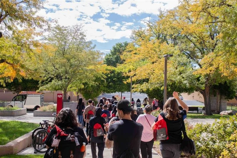 People walking in a park with trees showing fall colors, under a partly cloudy sky.