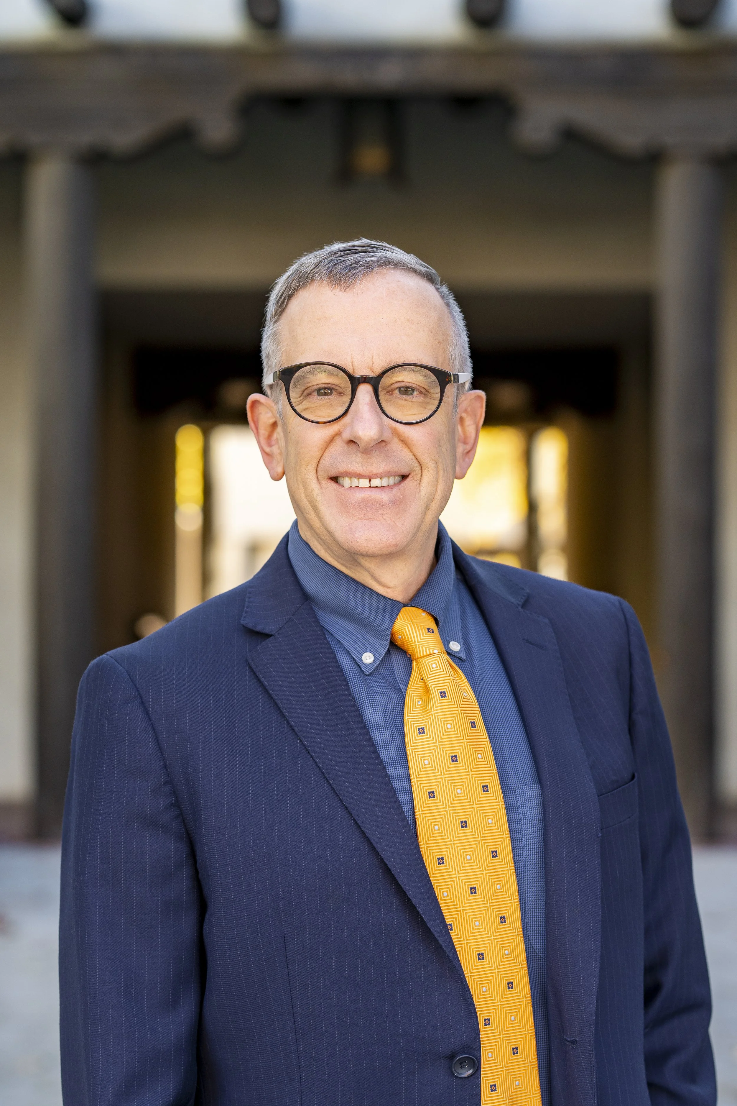 A man wearing glasses, a navy suit, and a yellow patterned tie standing outdoors with a structure behind him.