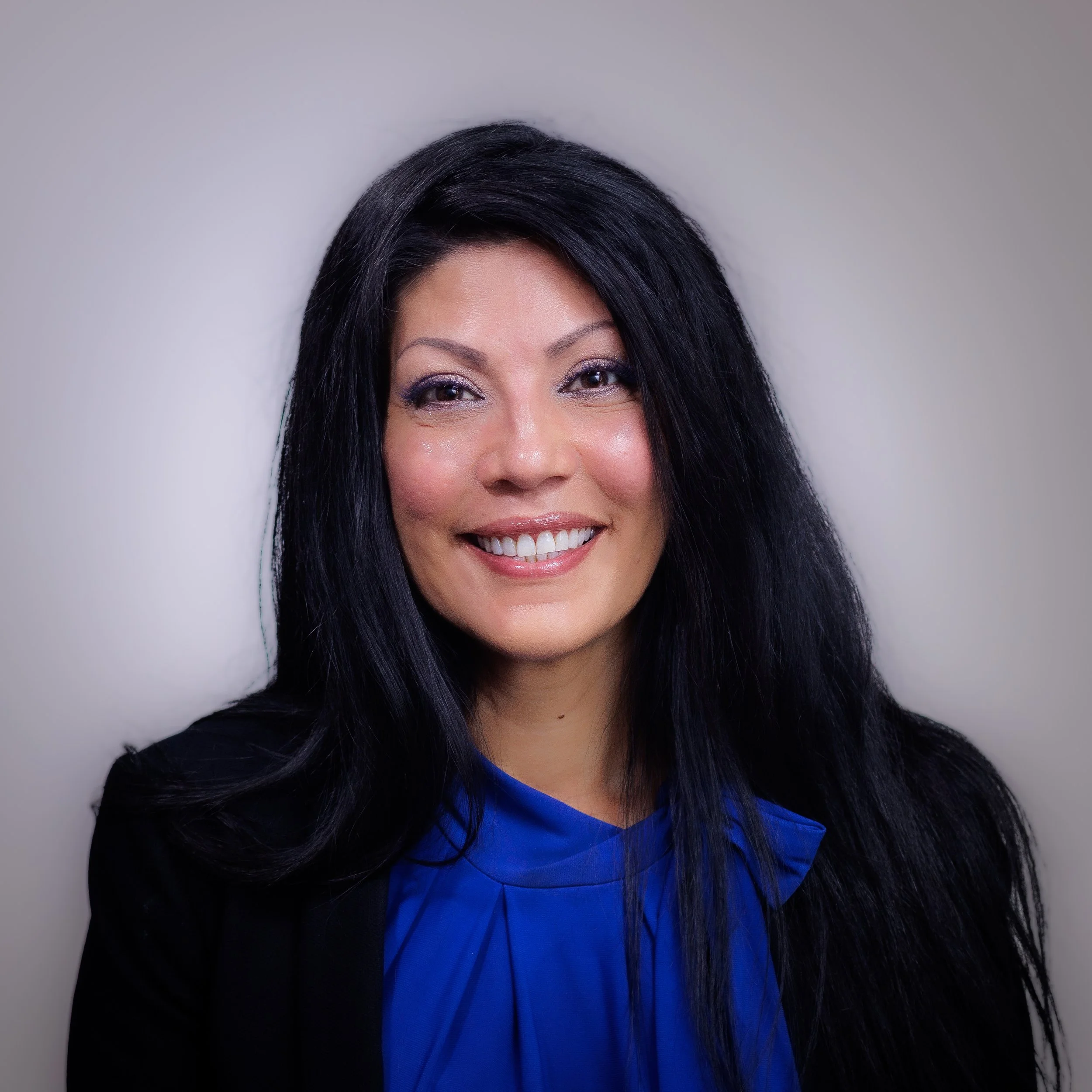 Partial view of a woman with black hair, makeup, and a bright smile, wearing a blue top and dark jacket, standing against a neutral background.