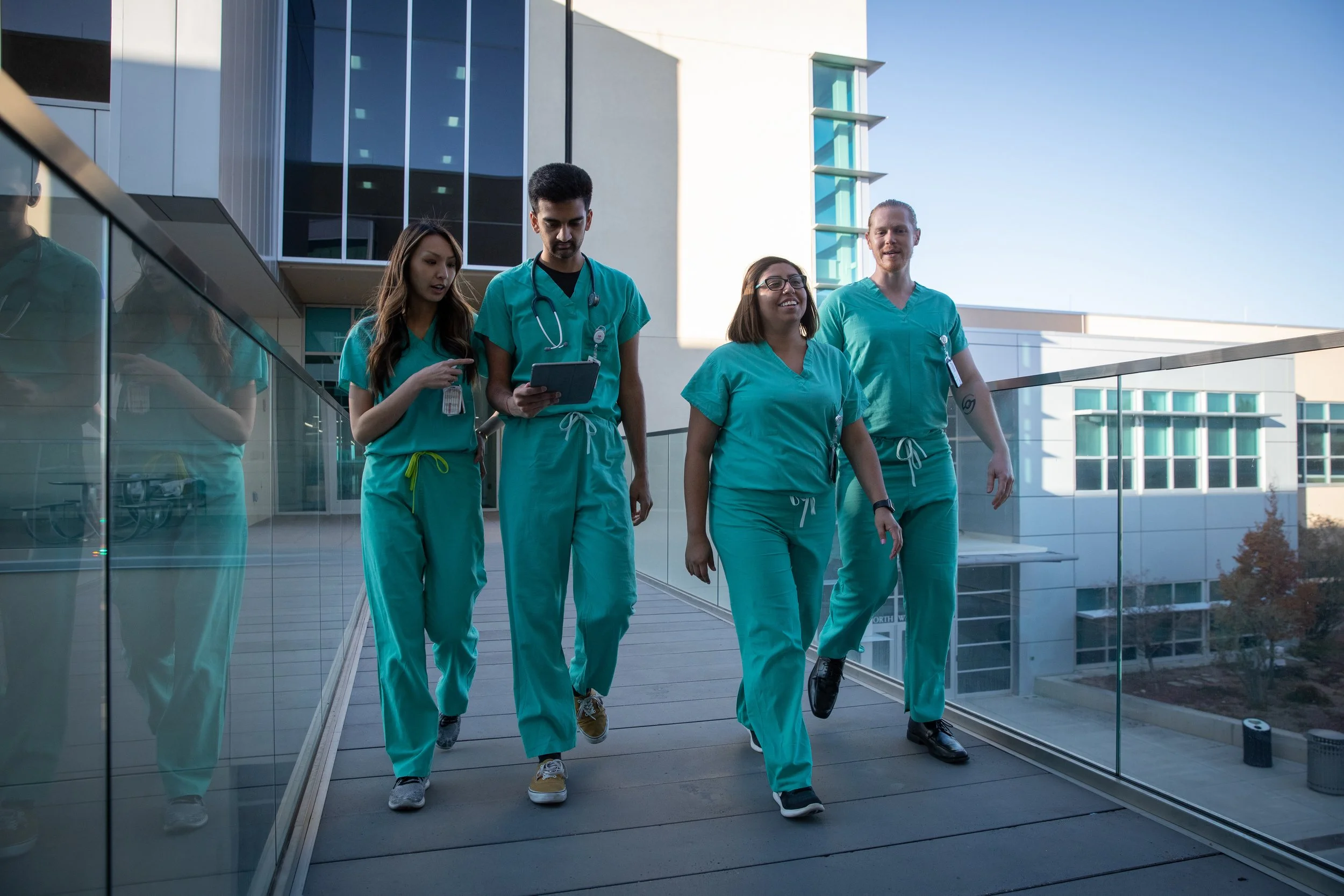 Group of four diverse healthcare professionals in scrubs walking outside a modern building during daytime, with one of them using a tablet device.