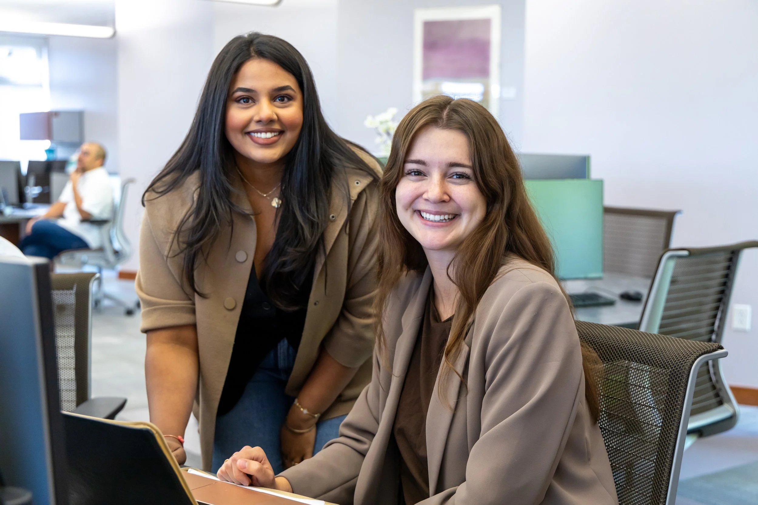 Two women smiling in an office, one standing and the other sitting at a desk with a computer monitor, with a man working at a desk in the background.