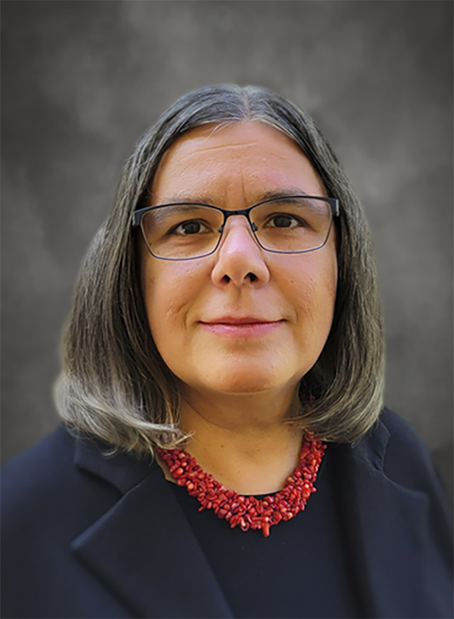 A woman with shoulder-length gray hair, glasses, wearing a black blazer and a red beaded necklace posing in front of a gray blurred background.