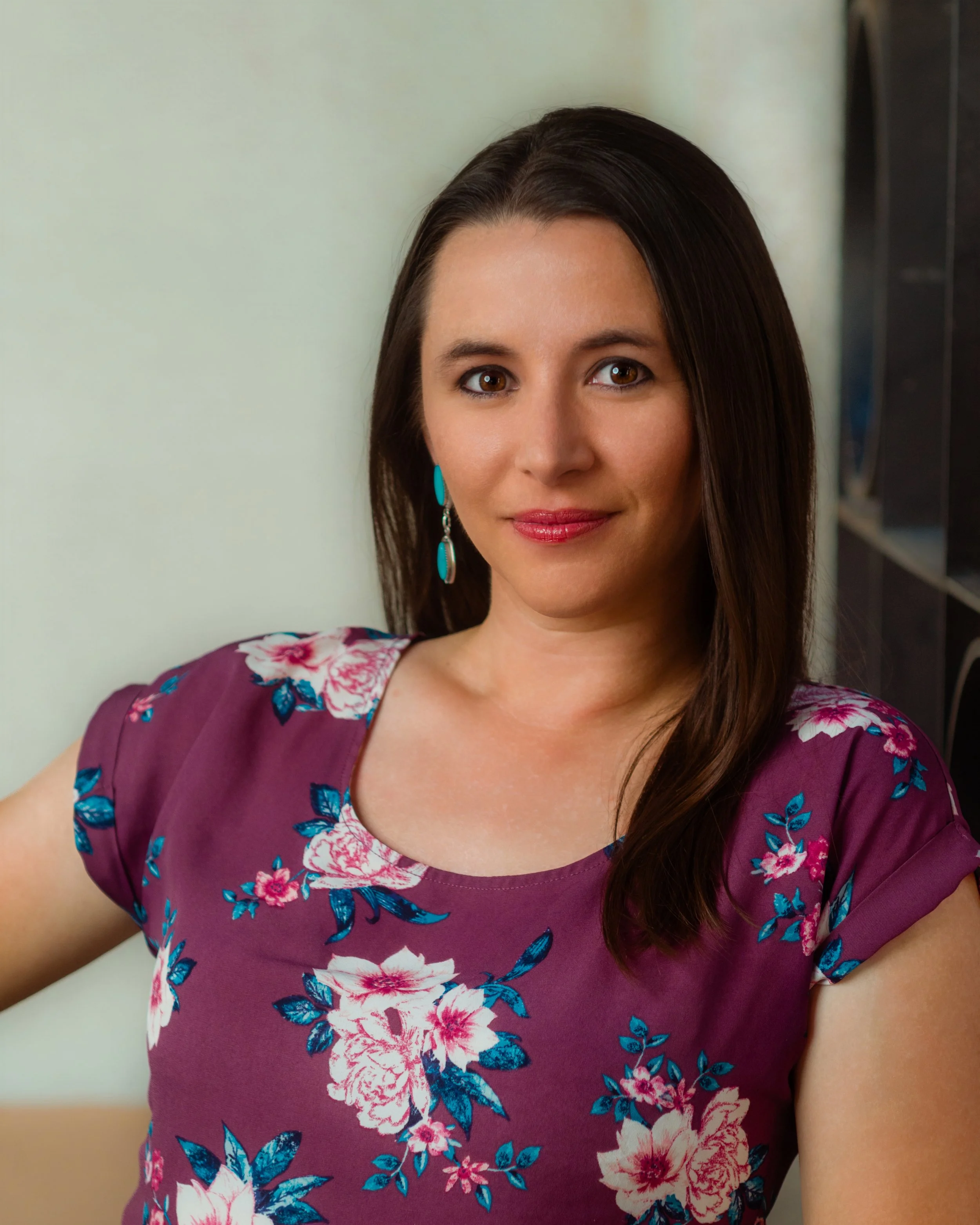 A woman with long dark hair and brown eyes, wearing a maroon dress with pink and blue floral pattern, standing indoors against a plain wall.