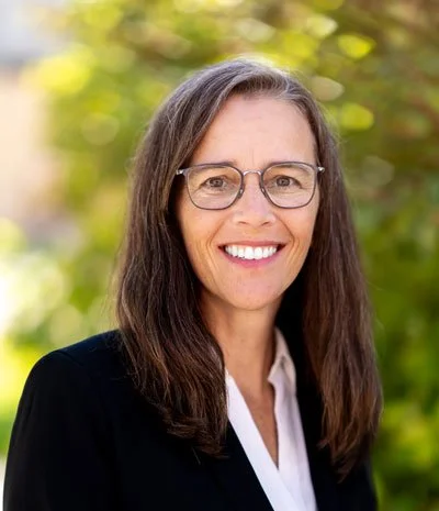 A woman with long brown hair and glasses smiling outdoors with green trees in the background.