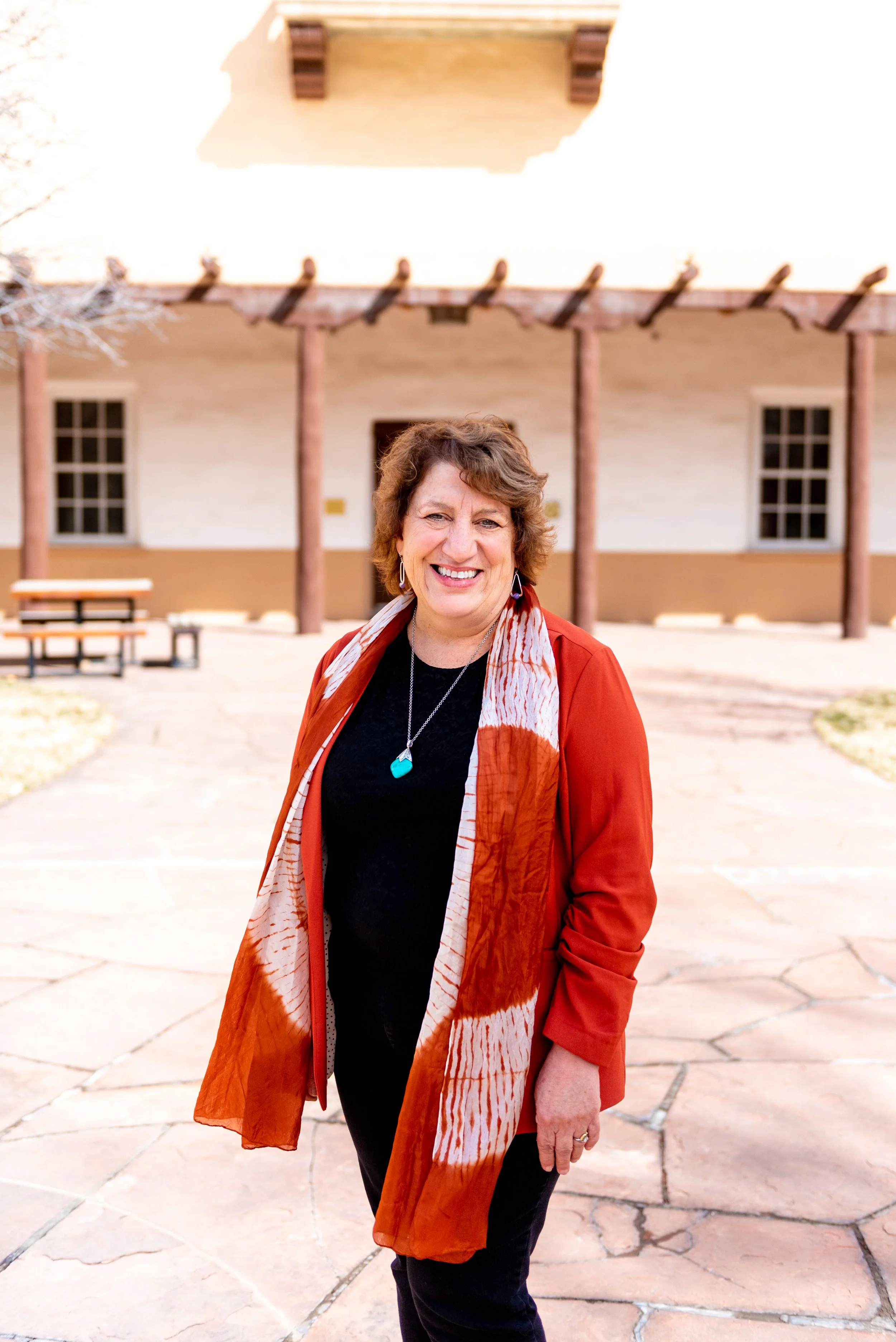 A woman in a burnt orange jacket and a patterned scarf standing outdoors in front of a light-colored building with columns. She is smiling and wearing a turquoise necklace.