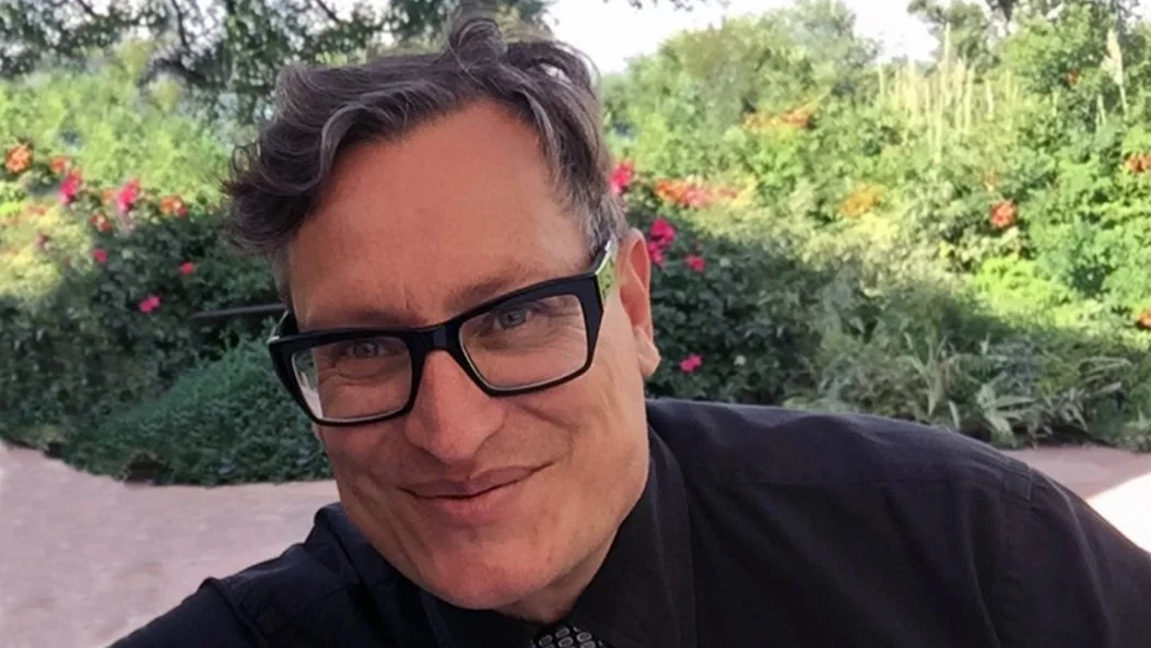 A man with glasses and short, wavy hair smiling outdoors with greenery and flowers in the background.