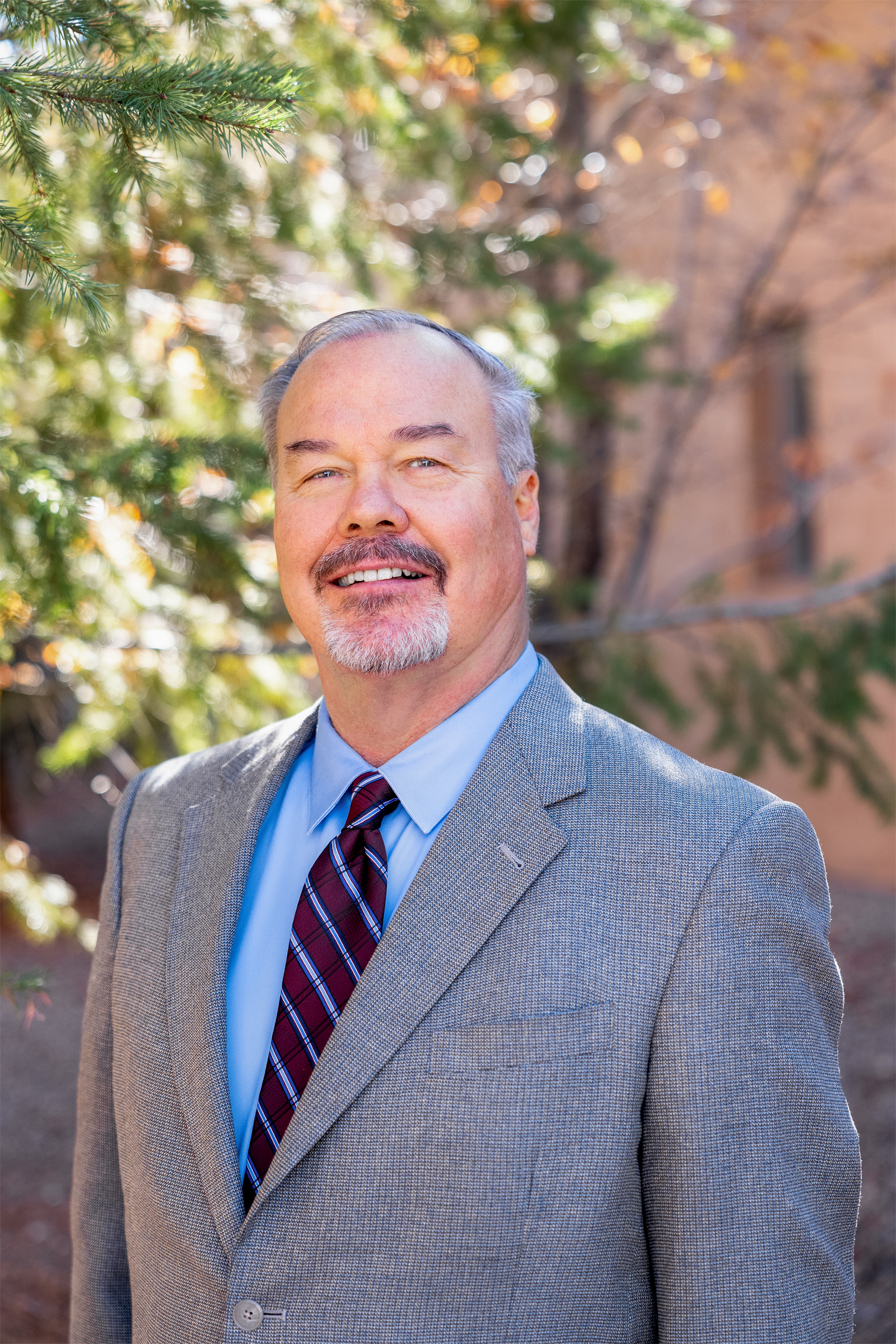 A middle-aged man with gray hair and a beard, wearing a gray suit jacket, light blue shirt, and a plaid tie, standing outdoors with trees and a brick building in the background.