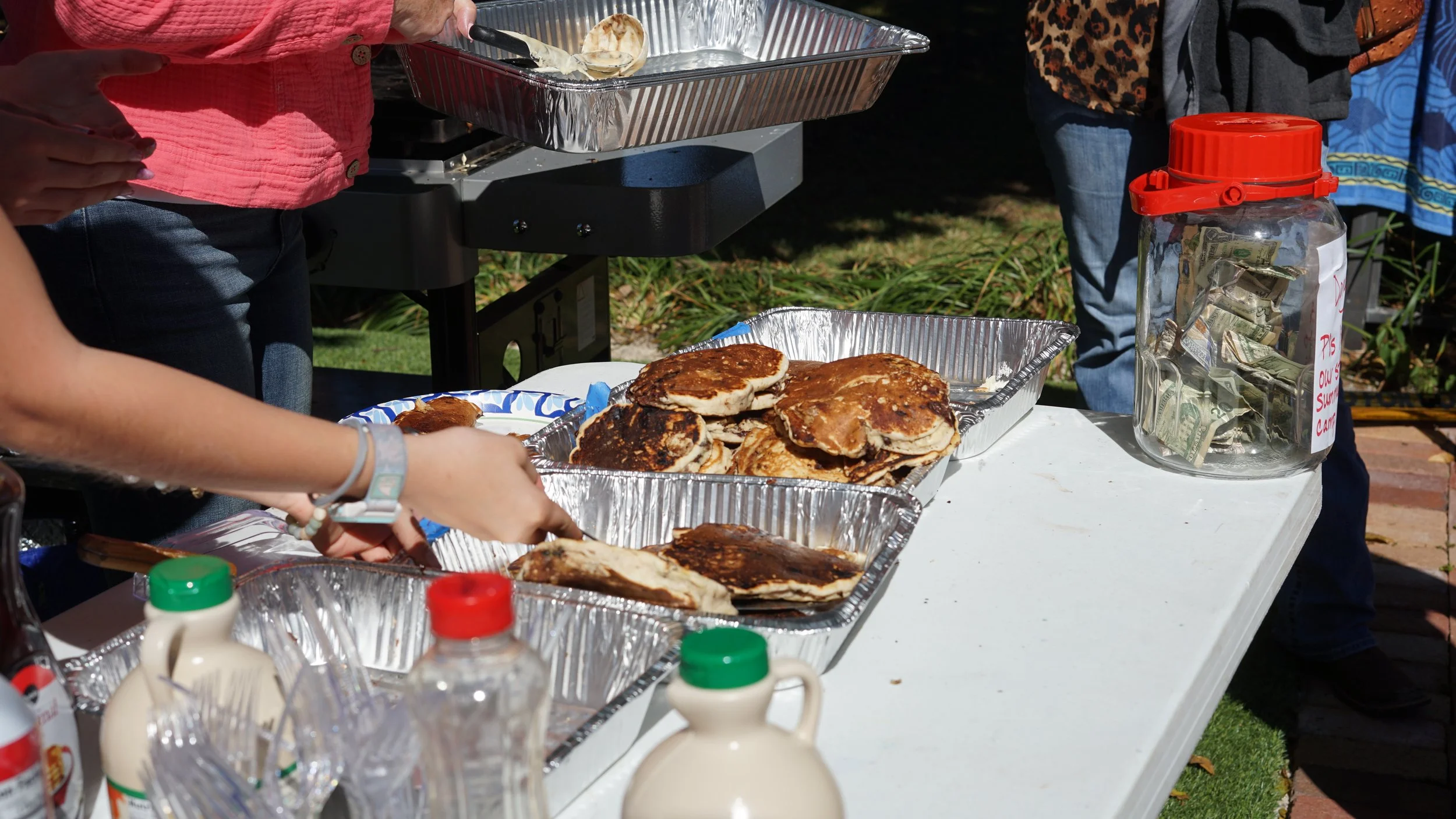 Tray of cooked pancakes on a table at an outdoor gathering, with a person serving food and a jar filled with cash in the background.