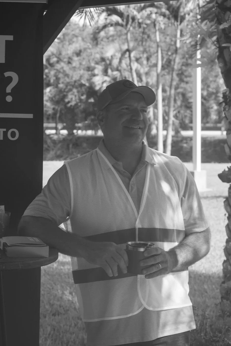 A smiling man in a cap and polo shirt holding a drink, standing outdoors near a vending area with trees in the background.