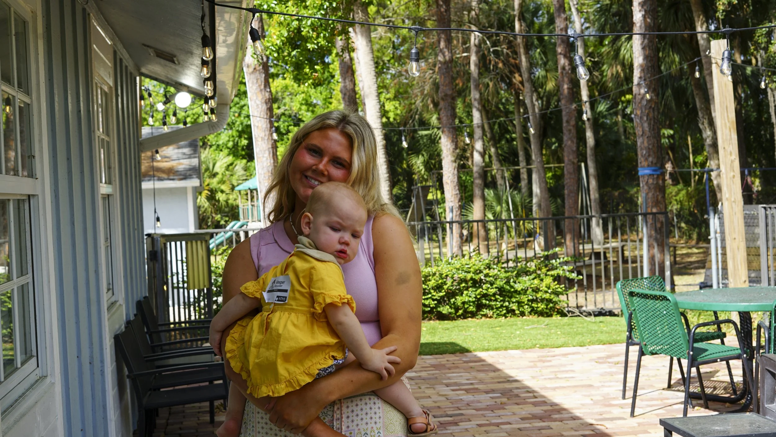 A woman holding a young child outdoors in a backyard with trees and patio furniture.