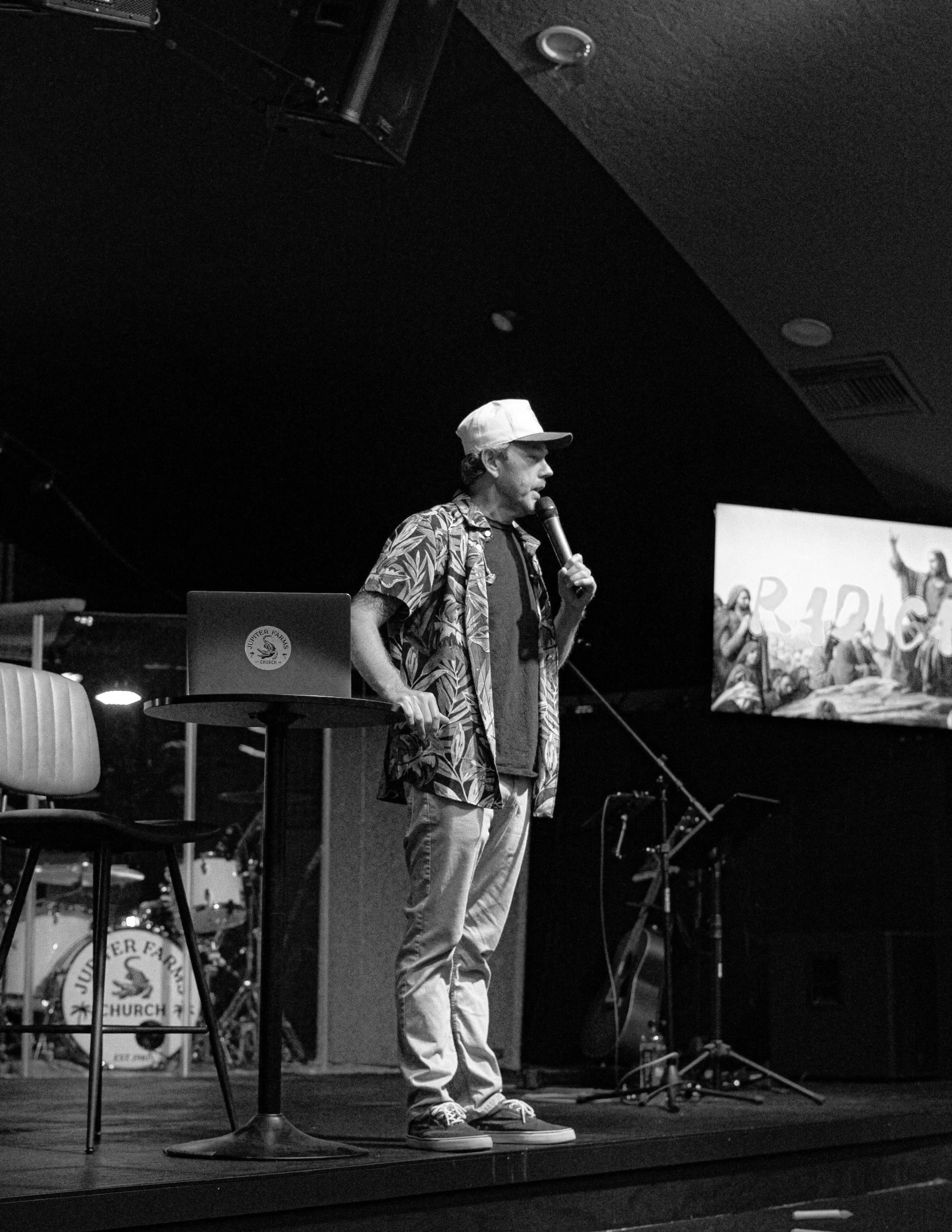 A man wearing a tropical shirt, baseball cap, and sneakers holding a microphone, standing on a stage with musical instruments and a screen in the background.