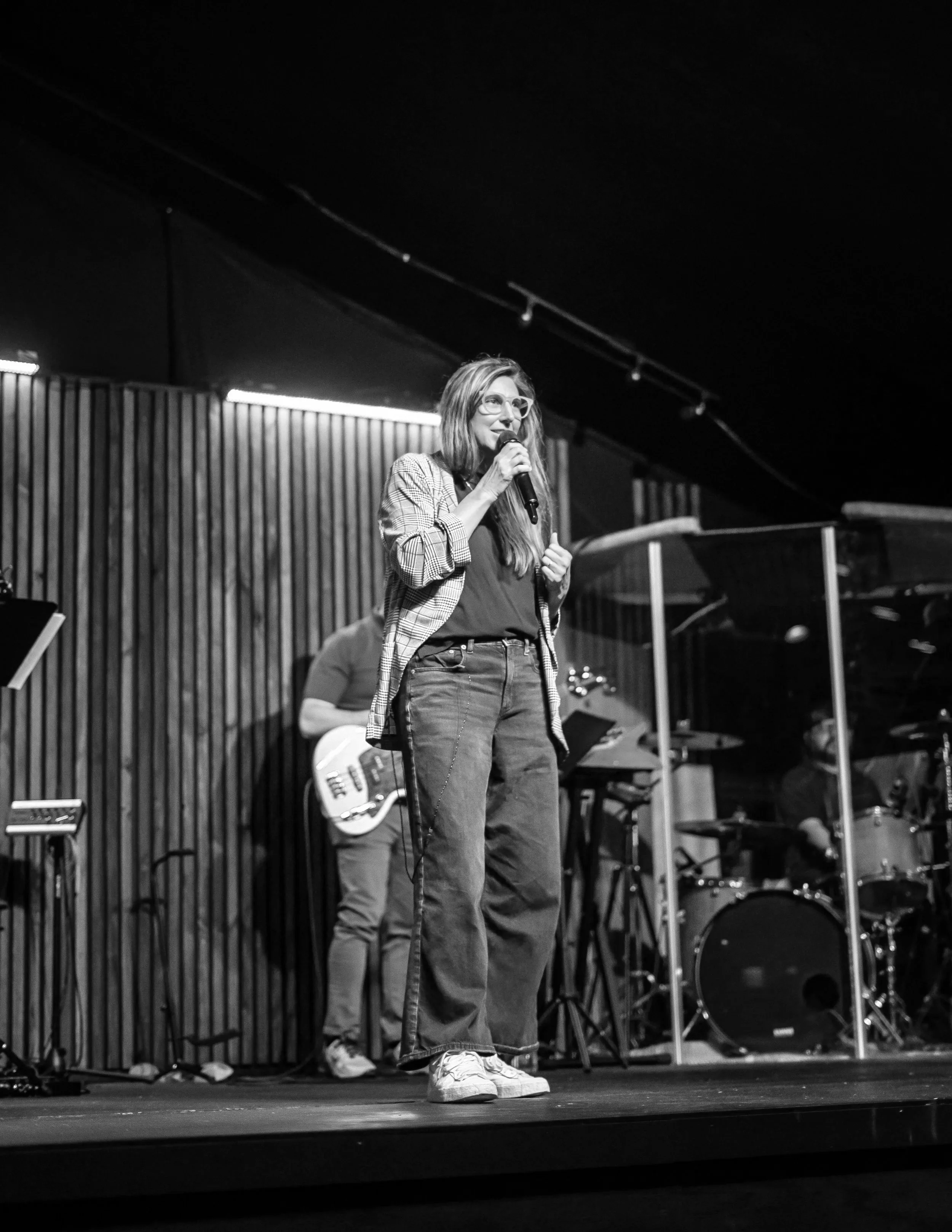 A woman with long hair, glasses, and a checkered blazer, holding a microphone and speaking on stage during a performance or speech. Behind her are a guitarist and drummer, with musical equipment and a wooden panel backdrop.