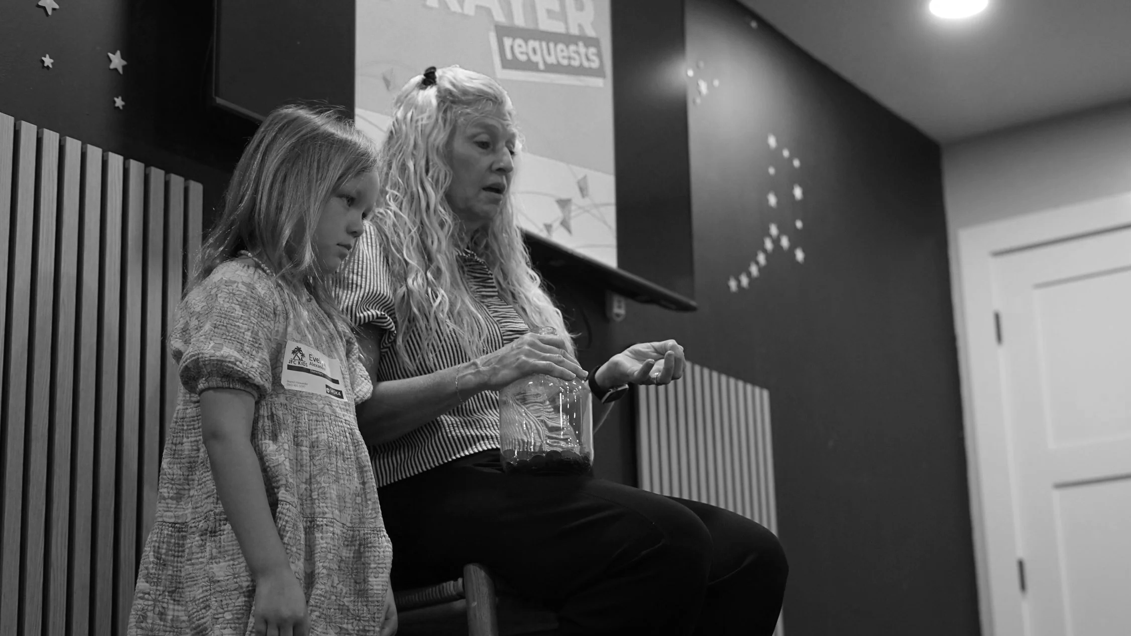 A woman and a young girl sitting on a stage, with the woman holding a plastic container filled with small objects. The setting appears to be an indoor event with a sign in the background that reads 'RAKER requests' and decorated with star-shaped stickers.