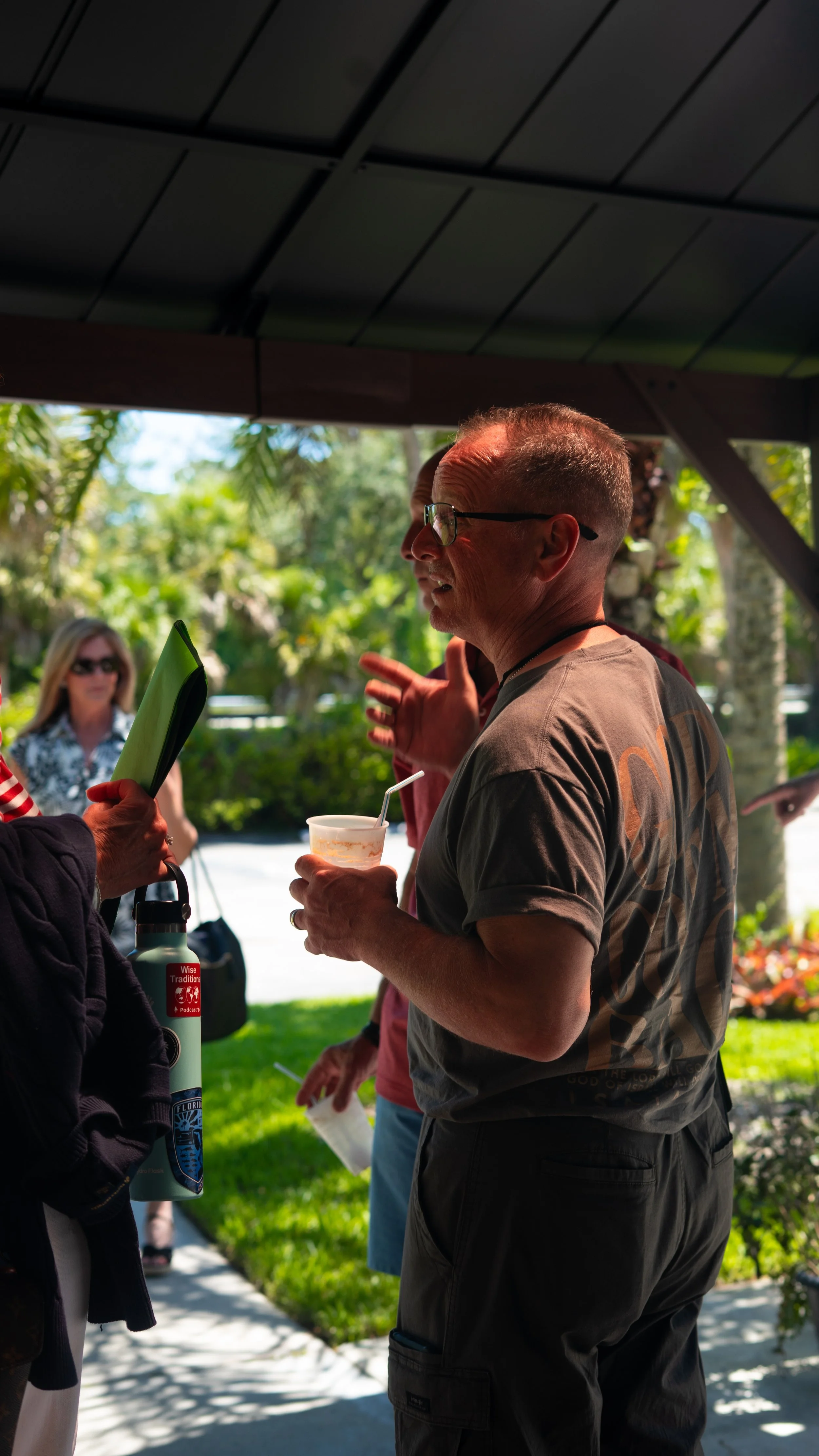 A man with glasses holding a drink and talking to a group of people outdoors under a shelter, with a woman holding a water bottle and a woman in the background wearing sunglasses.